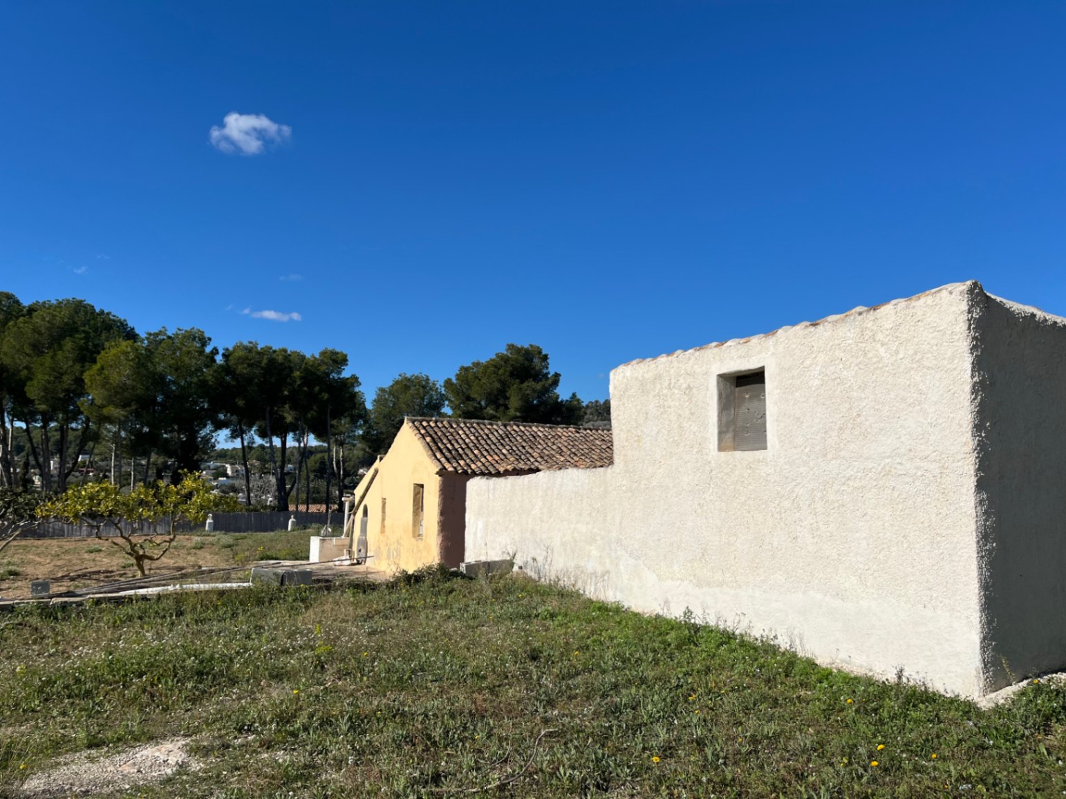 Vista exterior de la finca tradicional con muros de yeso rugoso en tonos blanco y ocre, techos de teja cerámica y parcela natural circundante.