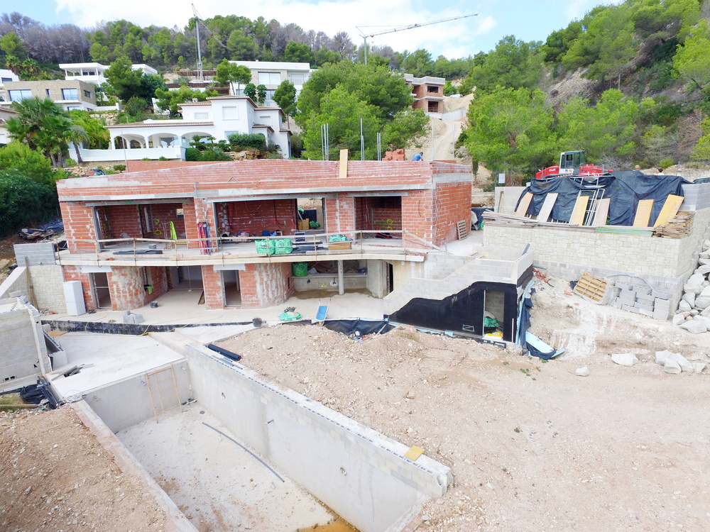 Fase de construcción de una villa de dos niveles con estructura de ladrillo visto y foso de piscina de hormigón en una parcela elevada rodeada de pinos.