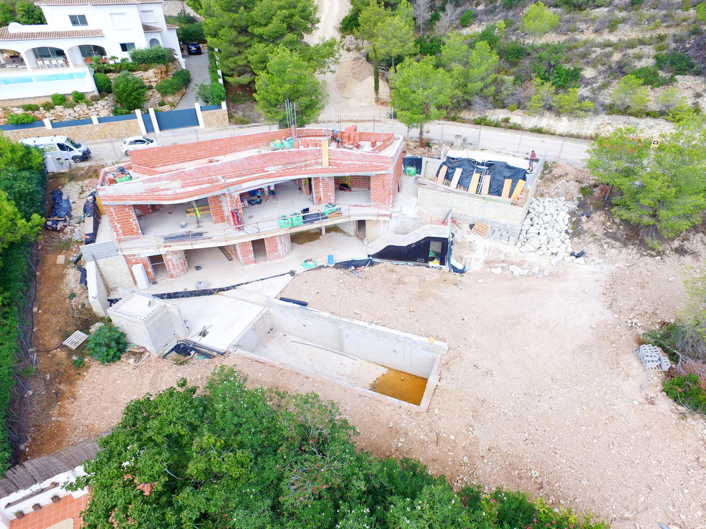Vista aérea de una villa en construcción con muros de ladrillo rojo, estructuras de hormigón y zona de piscina excavada en una parcela inclinada.