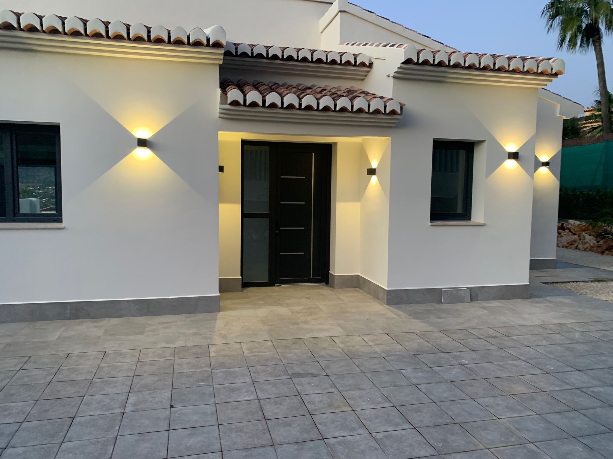 Entrada de villa moderna con puerta negra, fachada blanca y patio de gres gris. Apliques de pared simétricos aportan iluminación cálida bajo el tejado de teja tradicional.