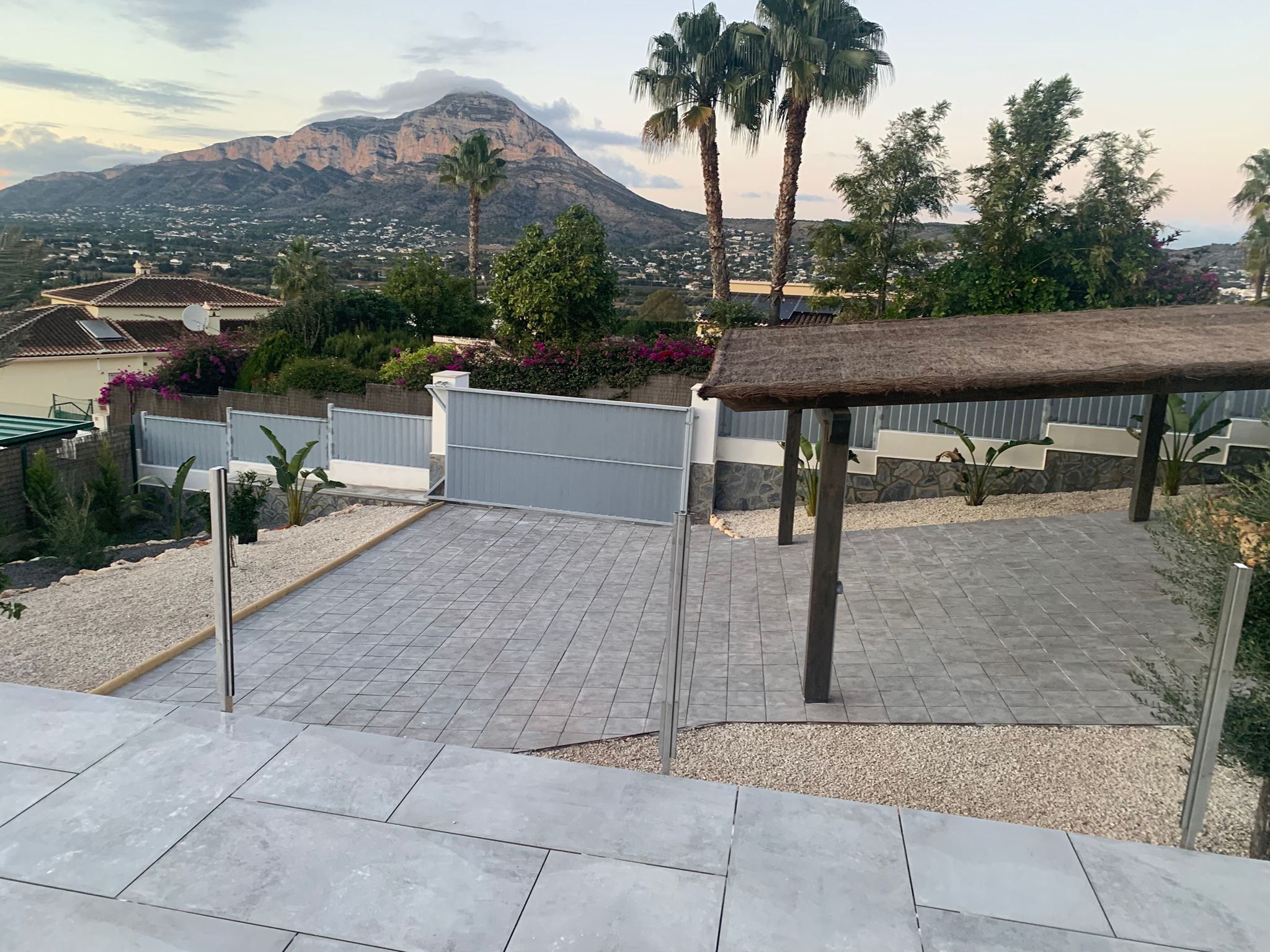 Entrada exterior pavimentada con baldosas grises, pérgola de madera con techo de brezo y vistas despejadas a la montaña bajo el cielo del atardecer.