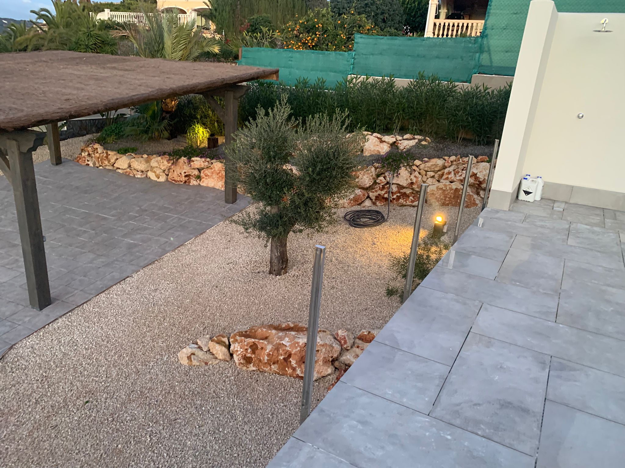 Jardín mediterráneo con zona de grava, un olivo joven, bordes de piedra natural, pérgola de brezo y terraza de gres gris con ducha exterior.