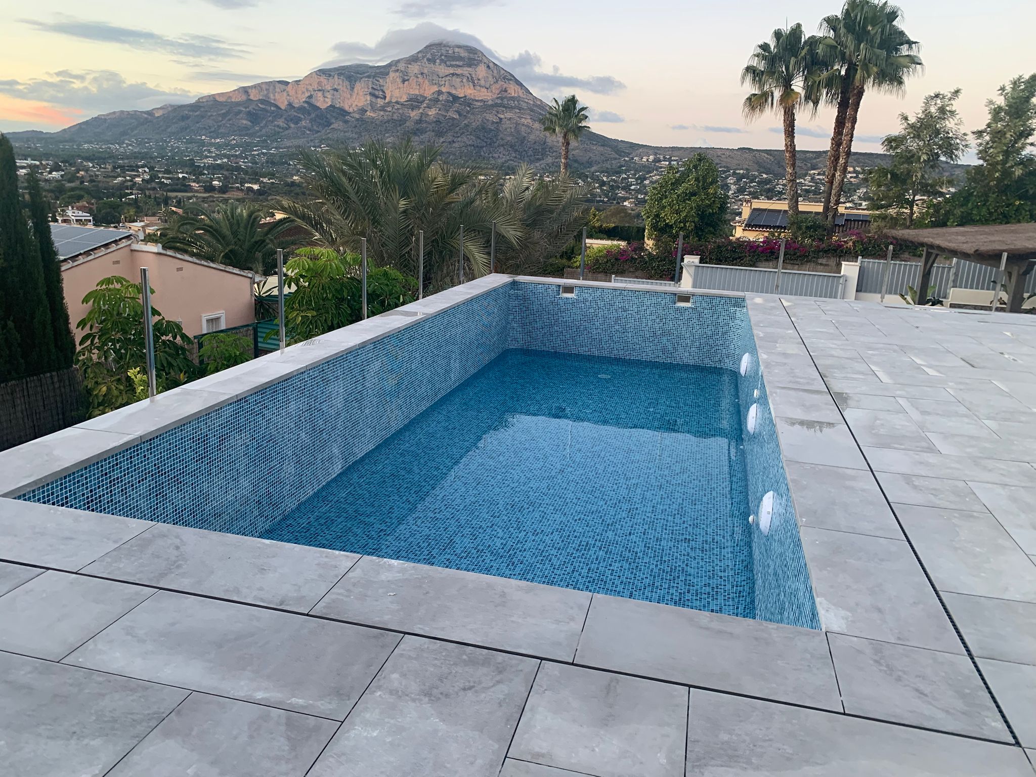 Piscina rectangular elevada con gresite azul y terraza de gres gris, protegida por barandillas de cristal con vistas panorámicas al monte Montgó.