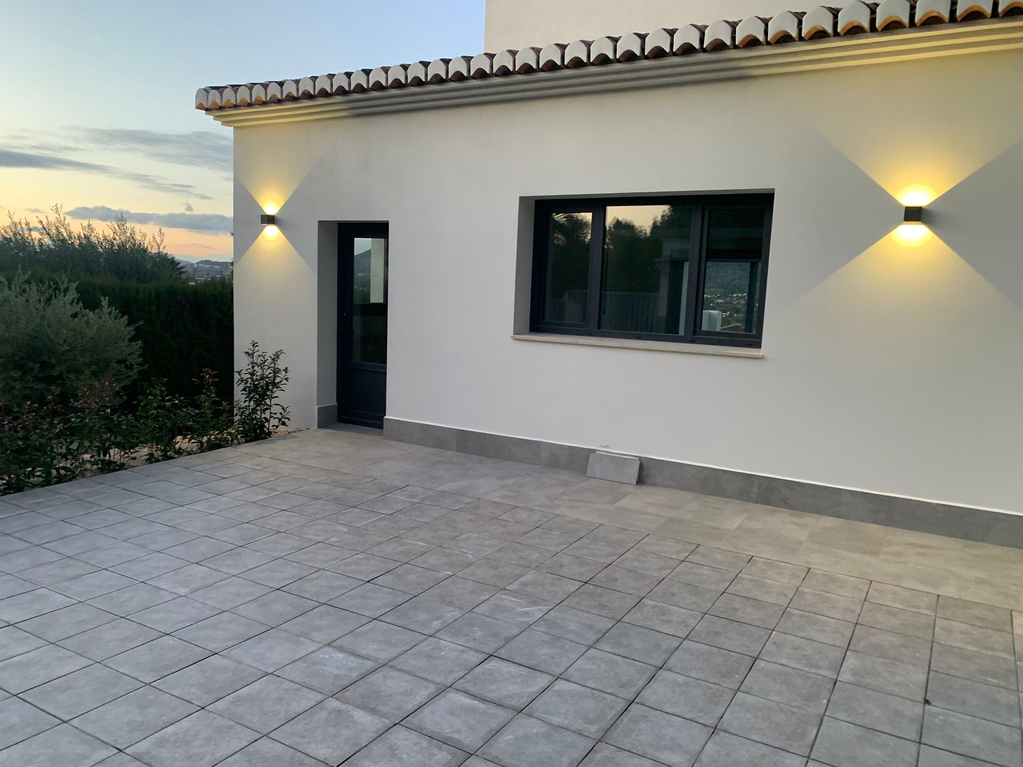Terraza exterior con pavimentación gris, fachada blanca e iluminación de pared bidireccional. Ventanales de carpintería oscura con vistas al paisaje montañoso al atardecer.