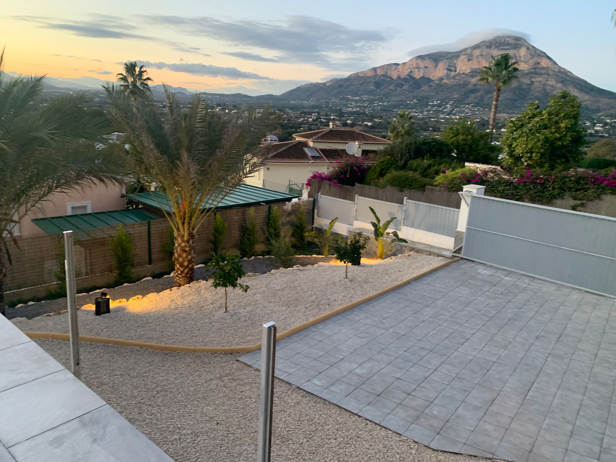 Vista elevada del jardín paisajístico con grava, palmeras y zona pavimentada, destacando las vistas panorámicas a la montaña durante el atardecer.