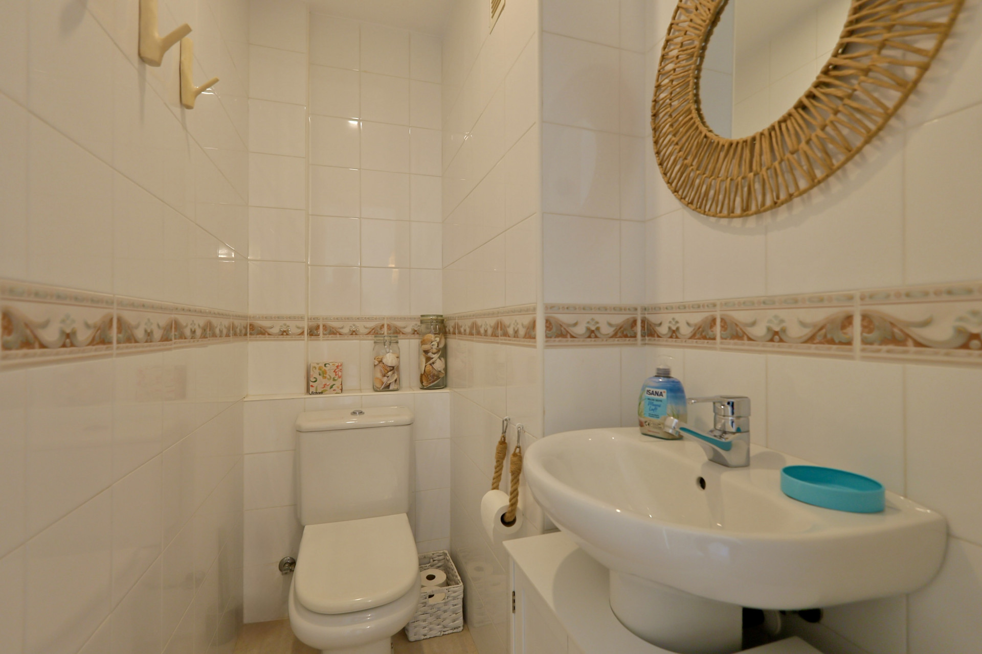 Aseo funcional con paredes alicatadas en blanco, cenefa ornamental, espejo de mimbre circular y lavabo compacto con grifería cromada.