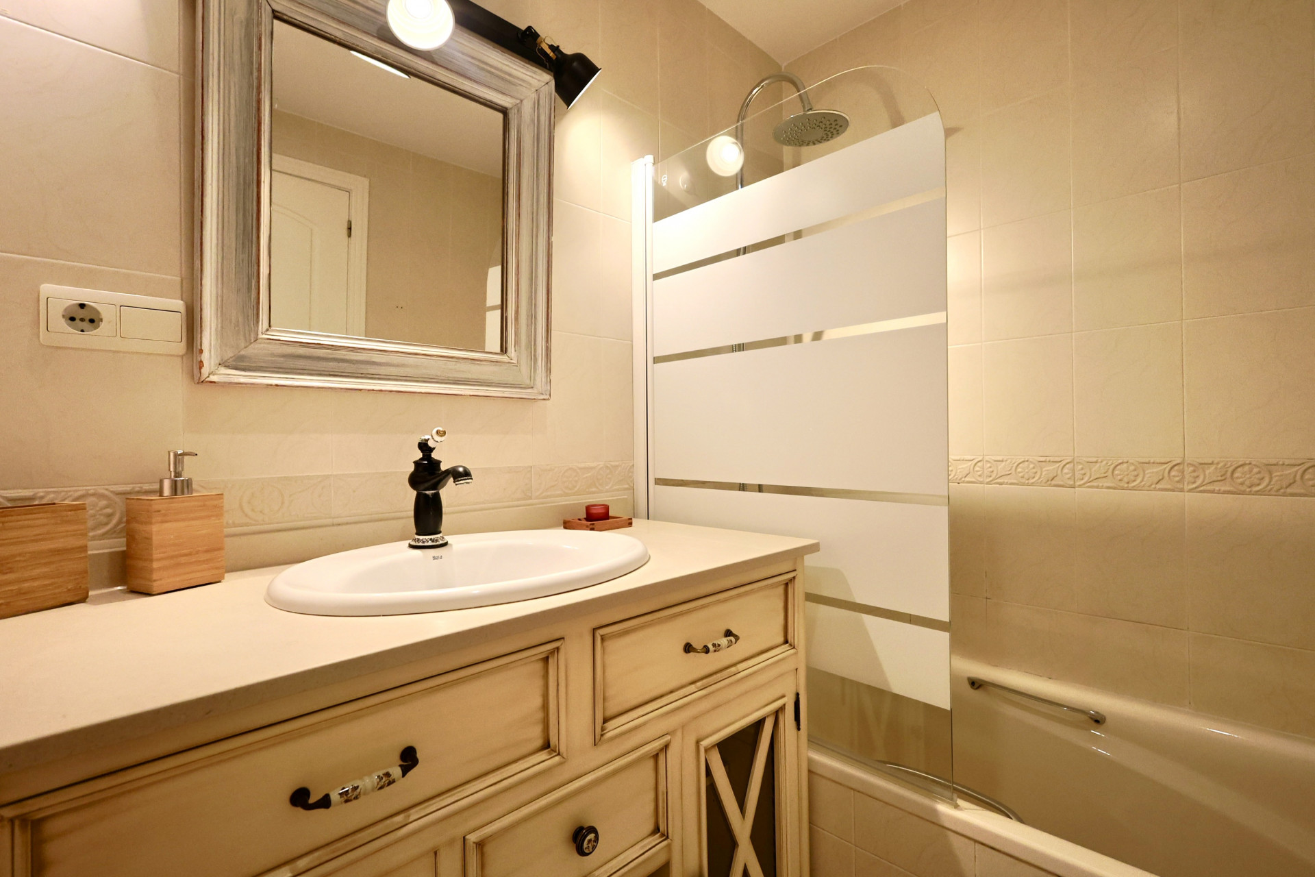 Baño con mueble de lavabo de estilo vintage, grifería negra, espejo enmarcado y bañera con mampara de cristal serigrafiado.