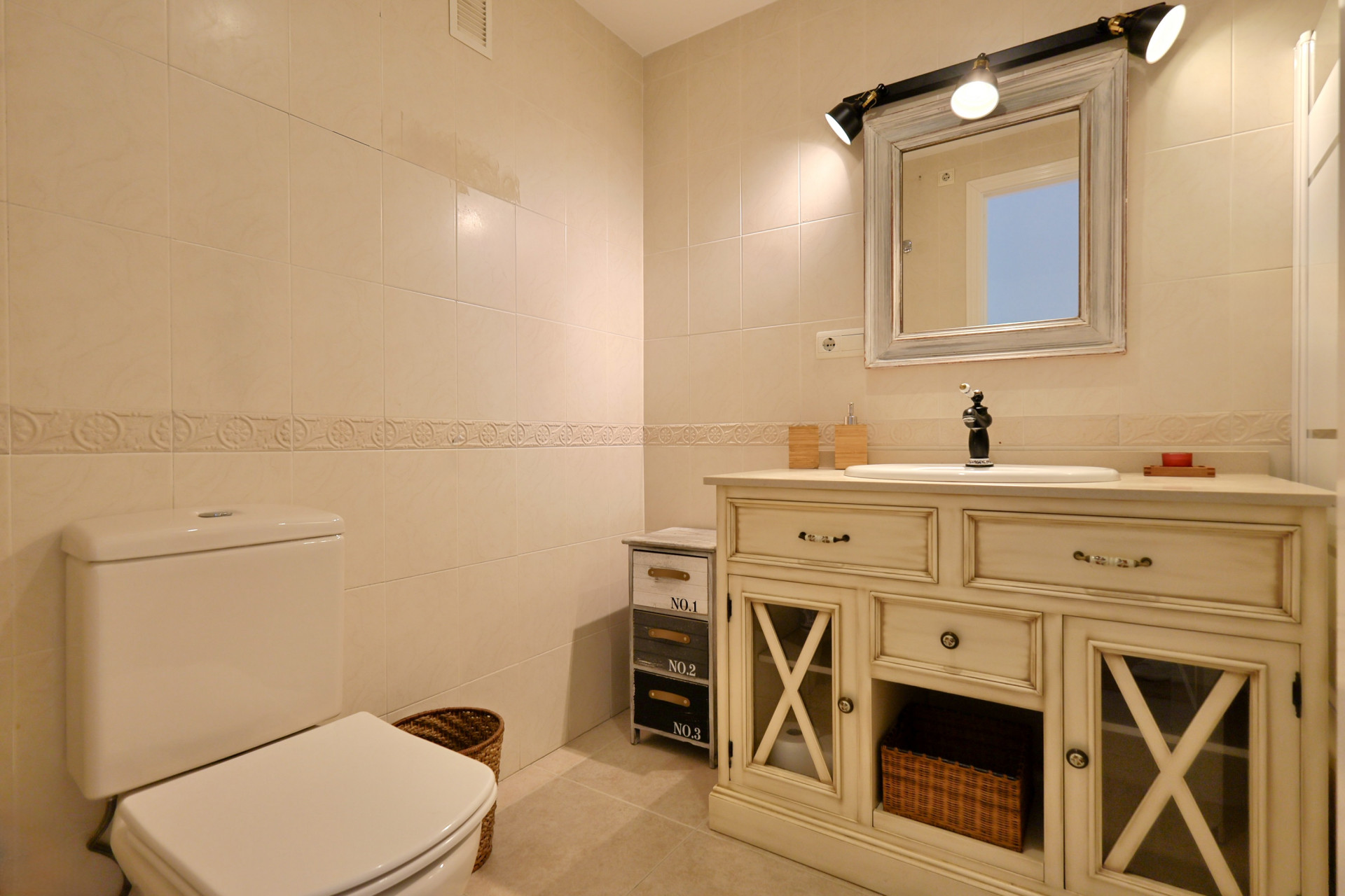 Baño con mueble lavabo de estilo vintage en color crema, puertas de cristal enrejadas, grifería negra y azulejos con cenefa decorativa.