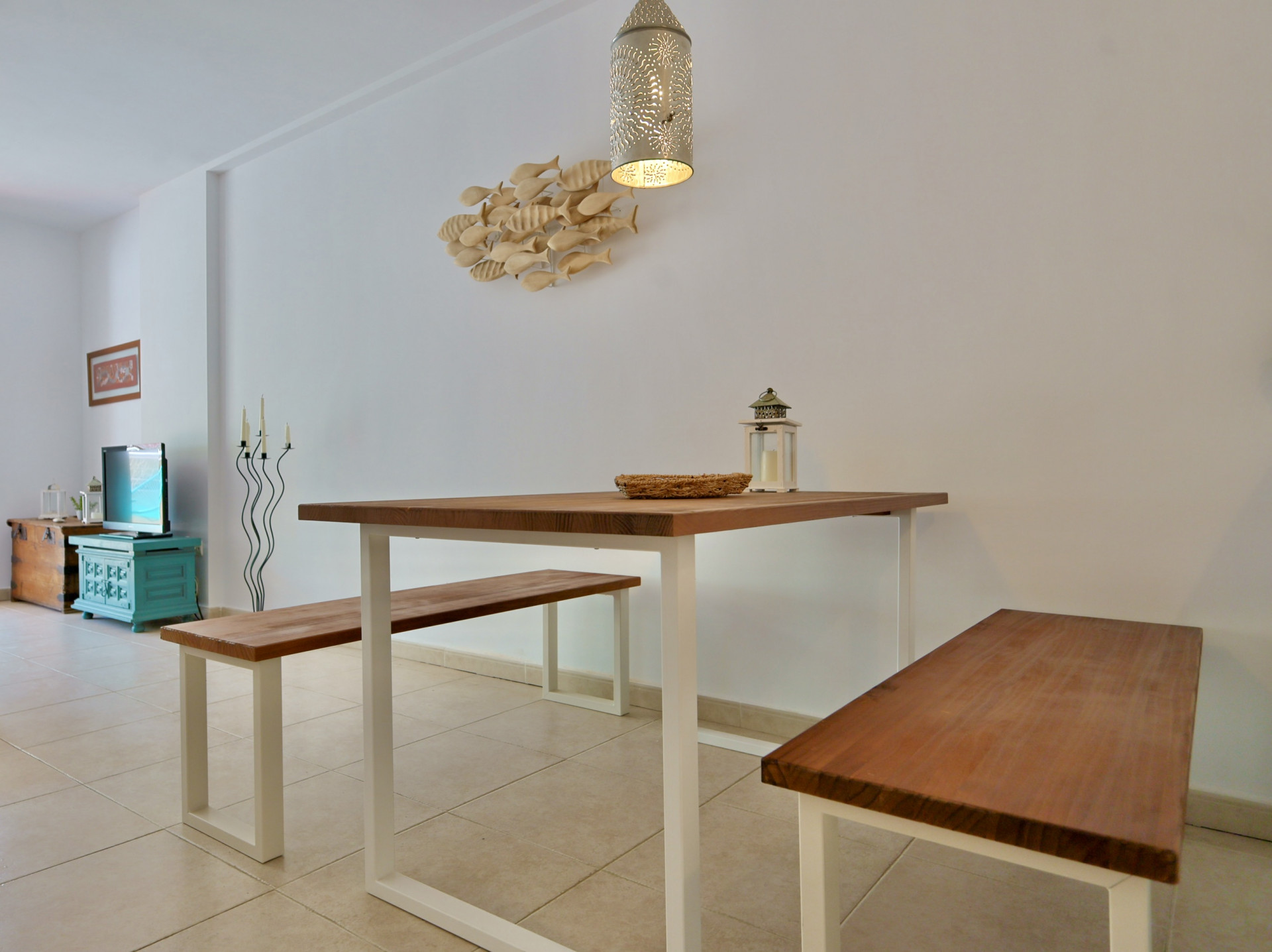 Comedor con mesa de madera maciza y bancos a juego sobre estructuras metálicas blancas, iluminado por una lámpara colgante perforada y decoración de pared náutica.
