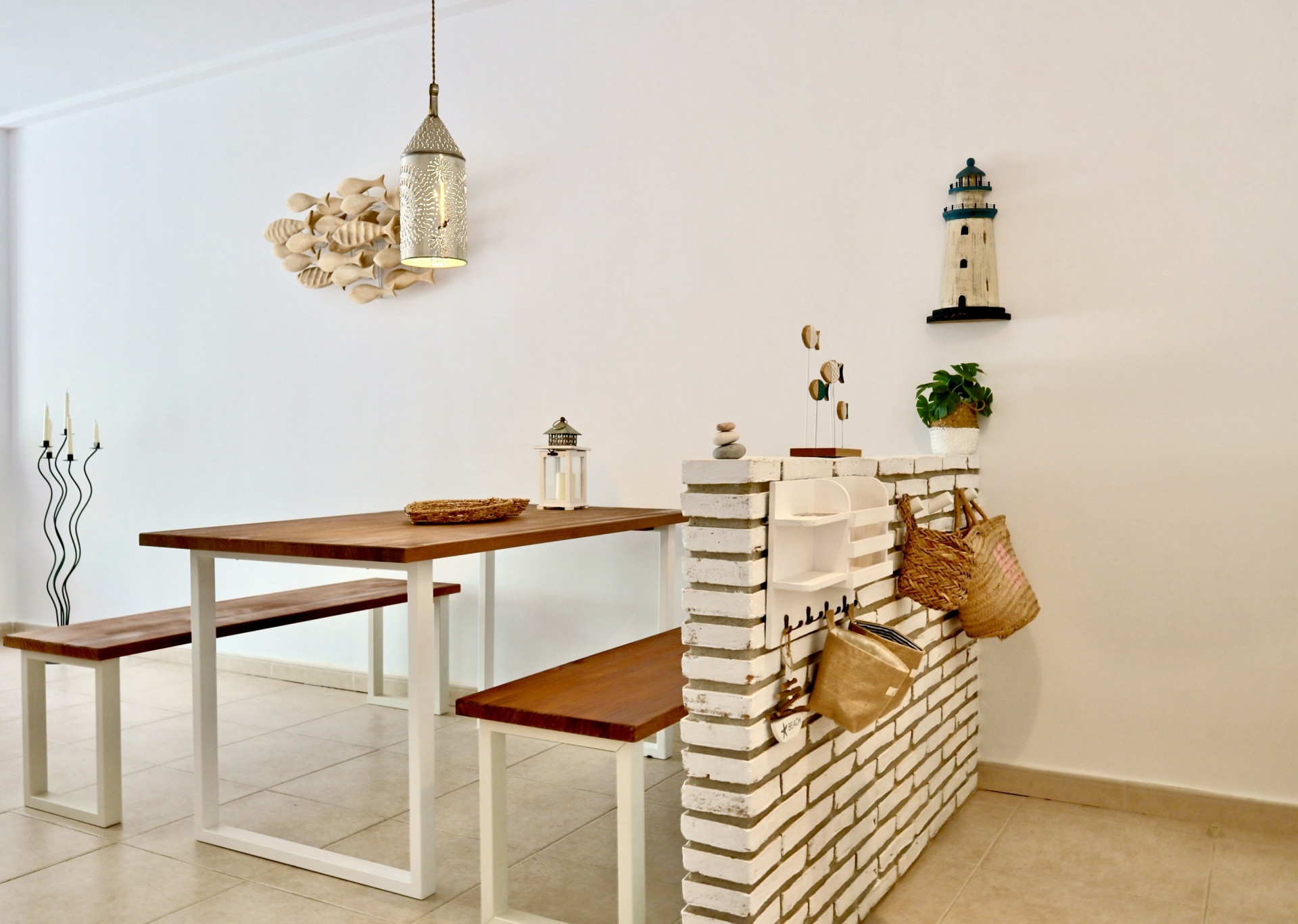 Comedor con mesa y bancos de madera sobre estructuras blancas, delimitado por un murete de ladrillo visto blanco y decoración de estilo marinero.