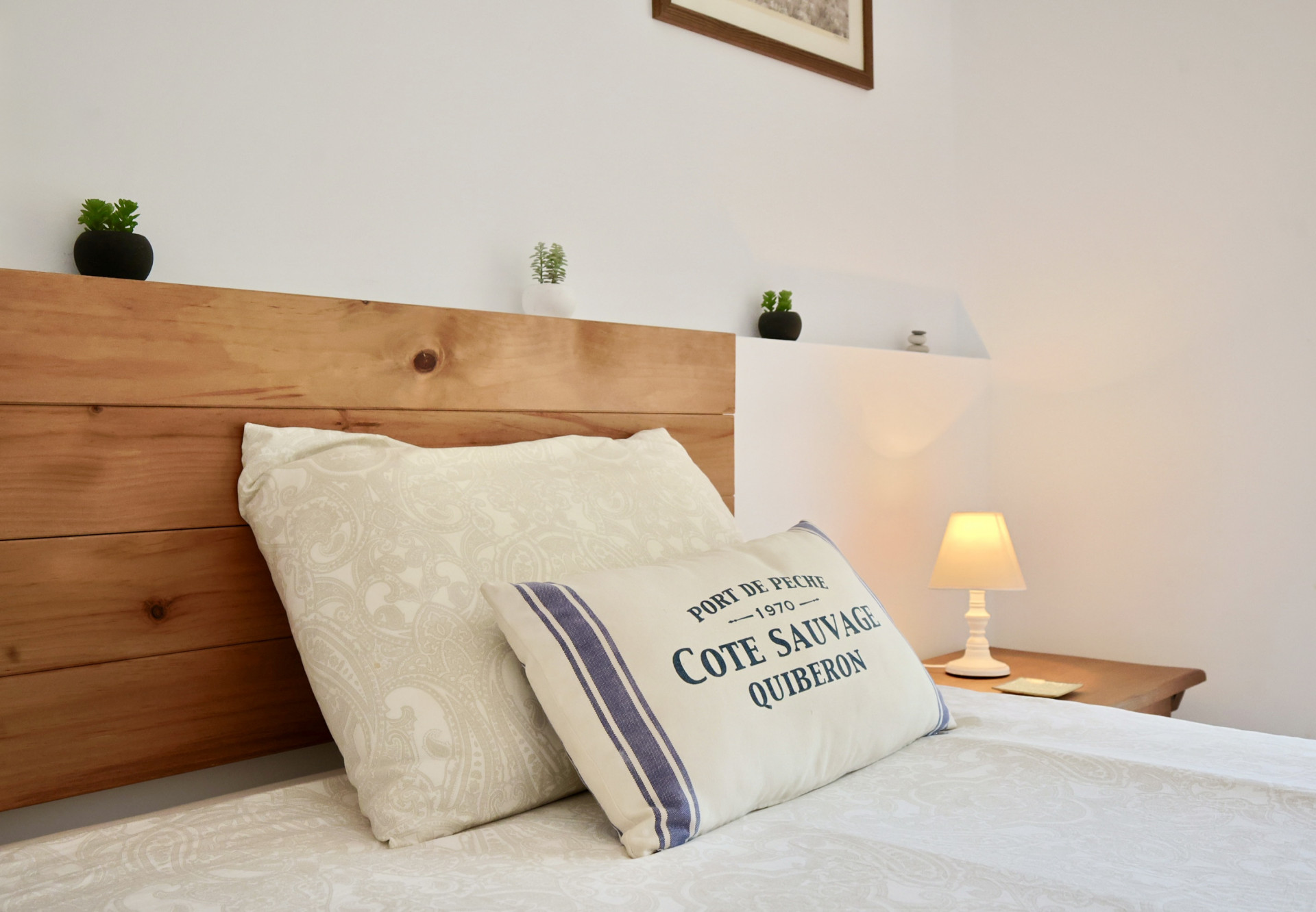 Detalle de dormitorio con cabecero de madera natural, cojines de estilo costero y una lámpara de mesa que aporta una iluminación cálida y acogedora.