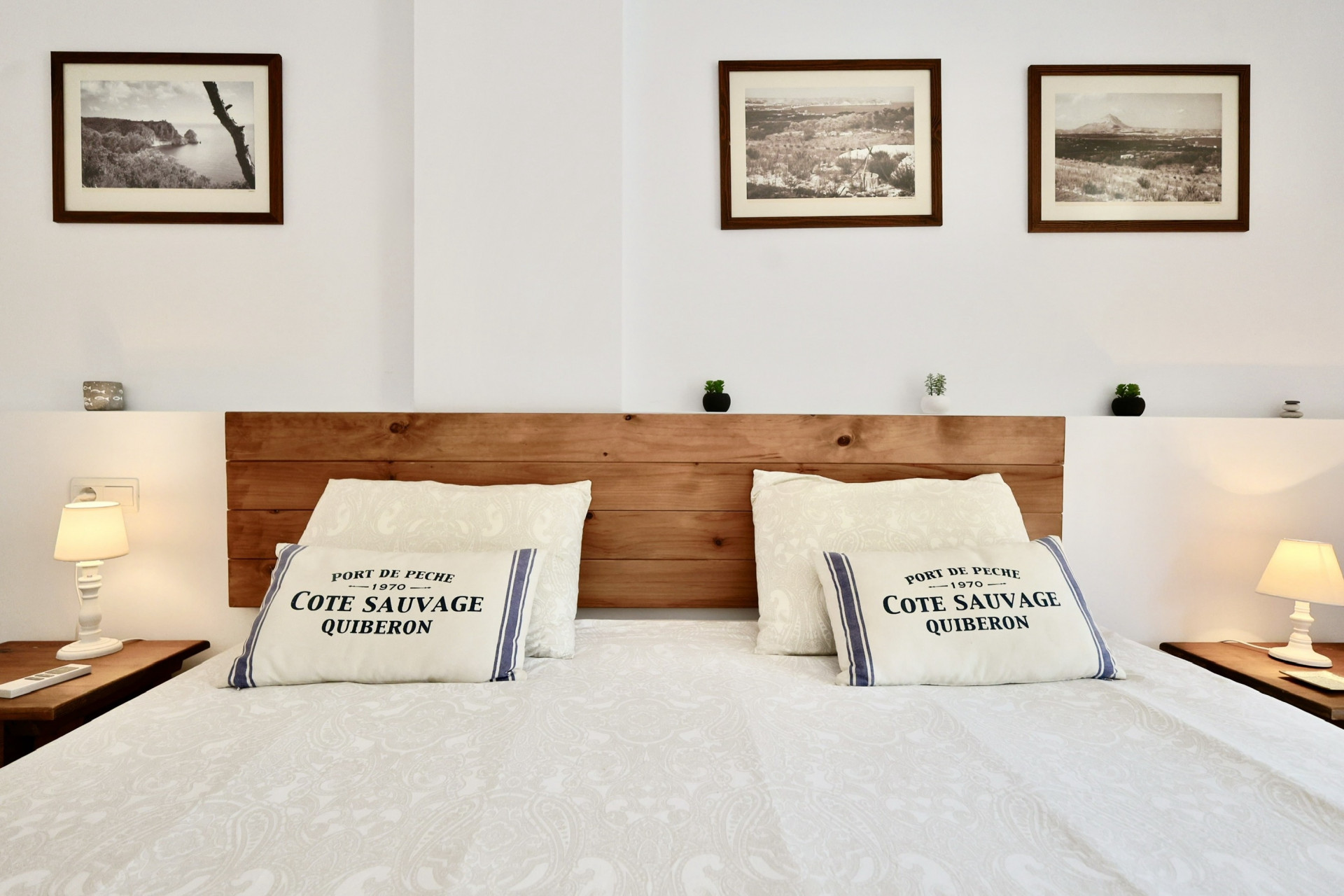 Dormitorio con cabecero de madera rústica, cojines de temática náutica y tres cuadros de paisajes. Incluye mesitas de noche simétricas con lámparas blancas de estilo clásico.