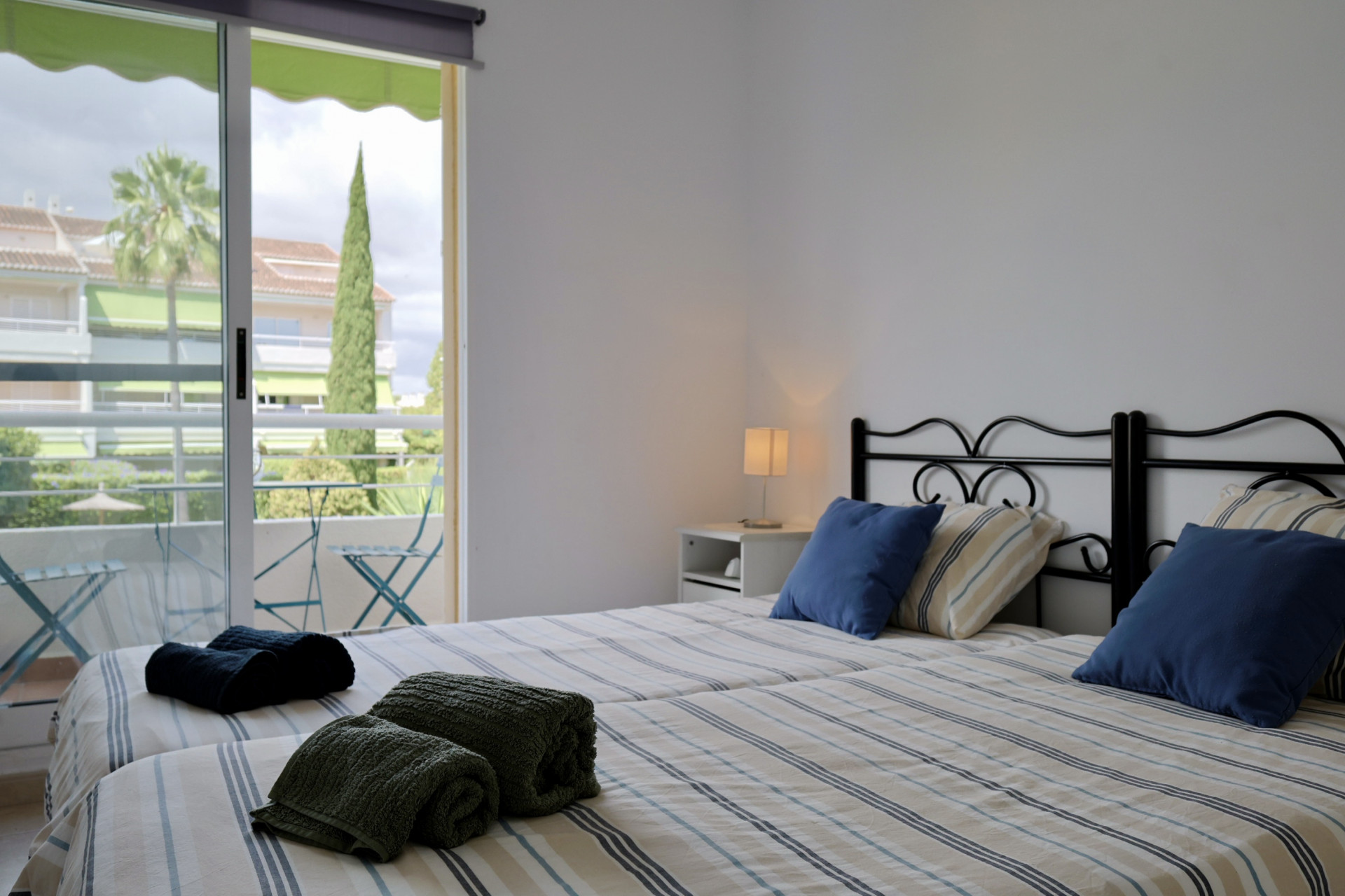 Dormitorio con camas individuales, cabeceros de forja negra y ropa de cama a rayas. Puertas correderas de cristal dan acceso a un balcón con vistas al jardín.
