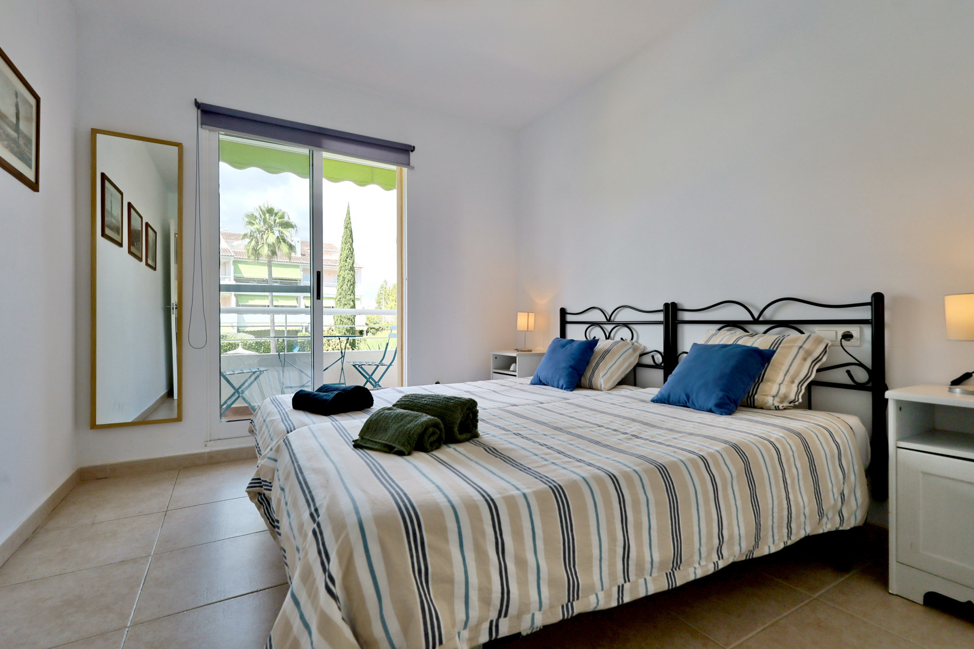 Dormitorio con camas individuales, cabeceros de hierro forjado negro y acceso directo a un balcón con vistas a zonas ajardinadas.