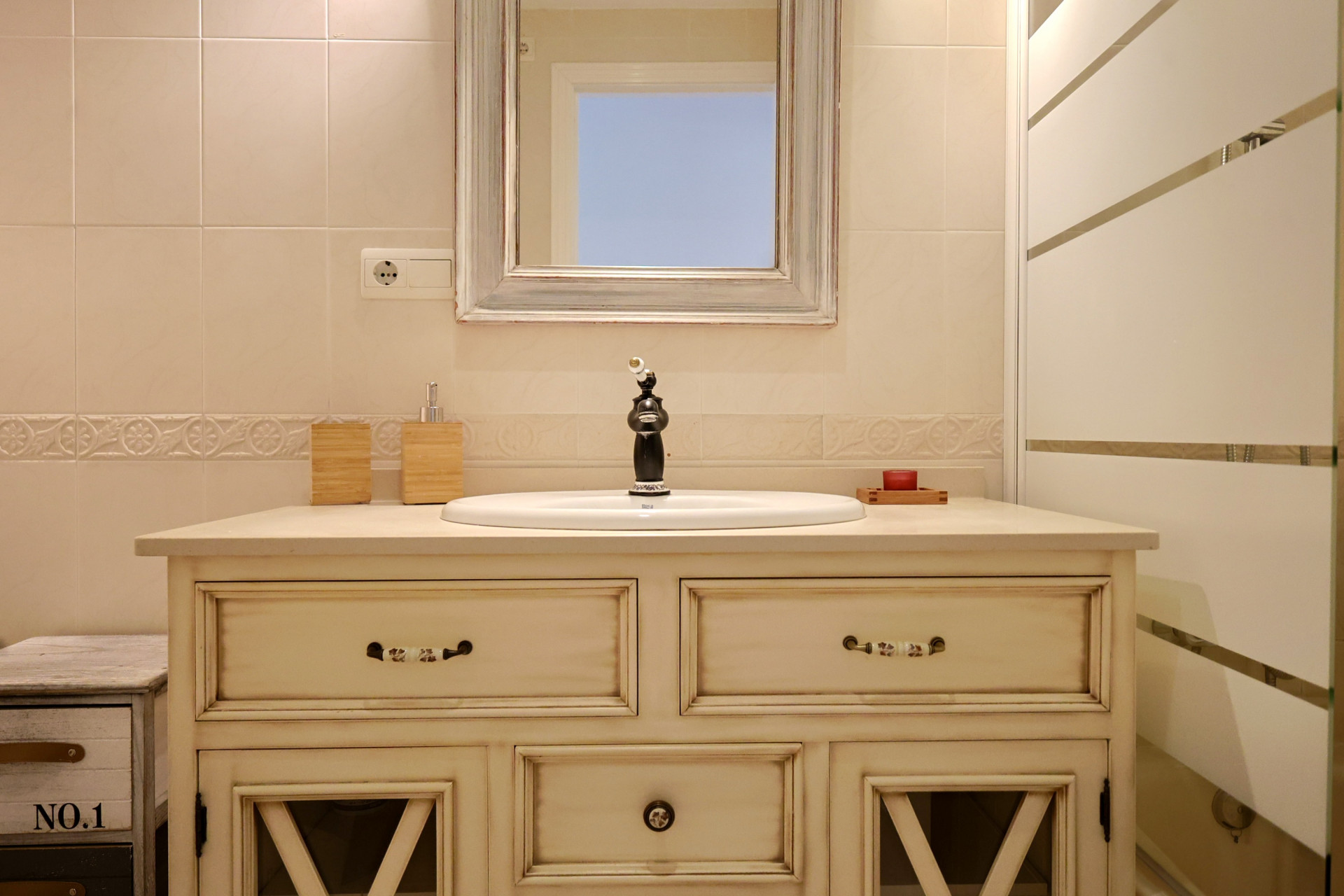 Mueble de lavabo de estilo vintage en color crema, con grifería negra ornamentada y un espejo de marco decapado sobre paredes con azulejos decorativos.