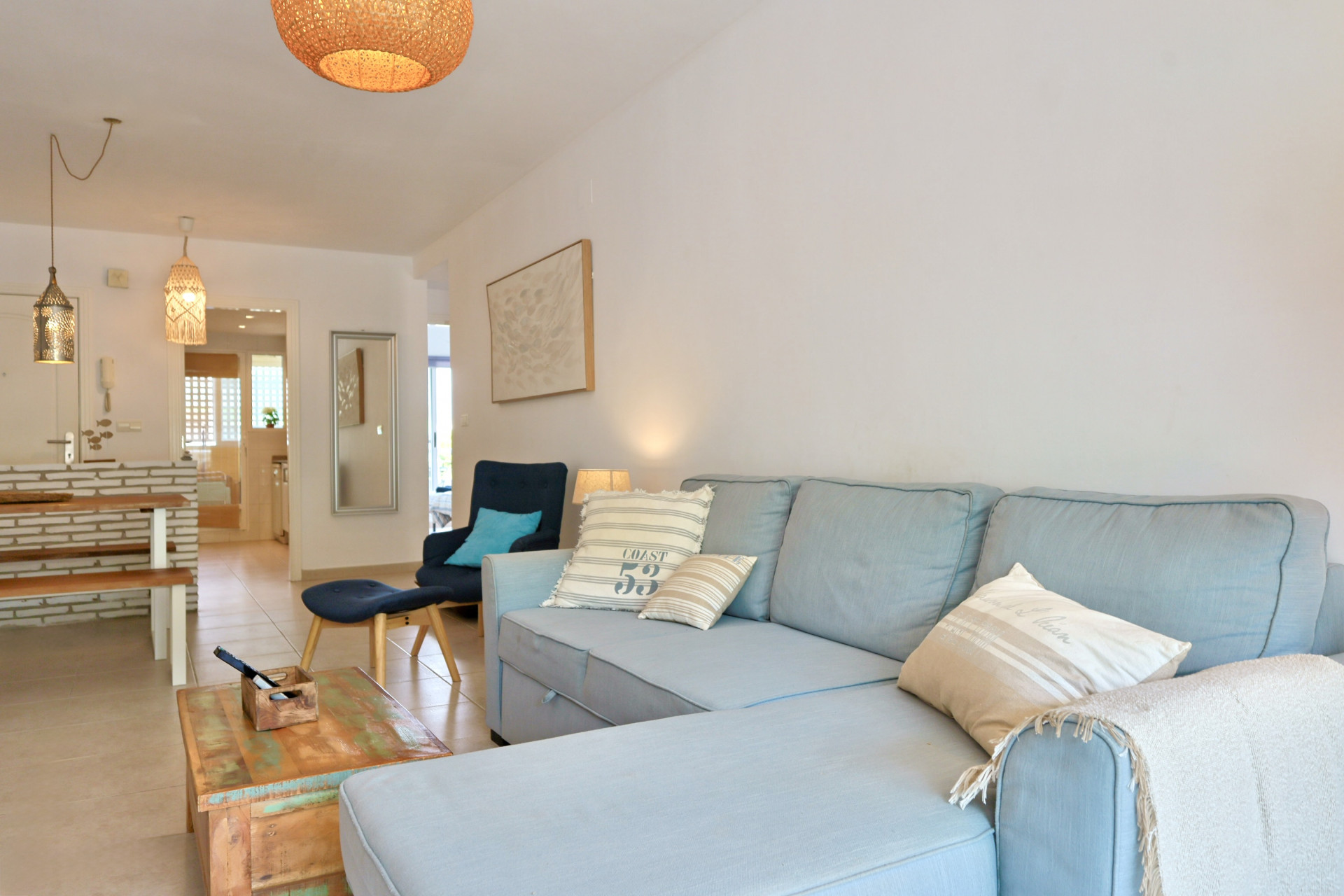 Salón de planta abierta con sofá rinconero azul claro, mesa de centro de madera rústica y zona de comedor con detalles en ladrillo y lámparas de mimbre.