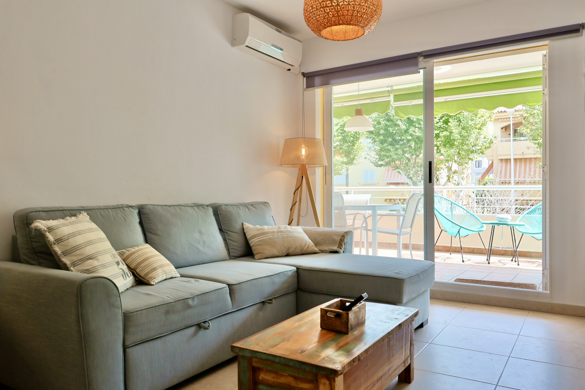 Salón luminoso con sofá rinconero gris, mesa de madera rústica y acceso directo a una terraza amueblada con sillas Acapulco turquesas.