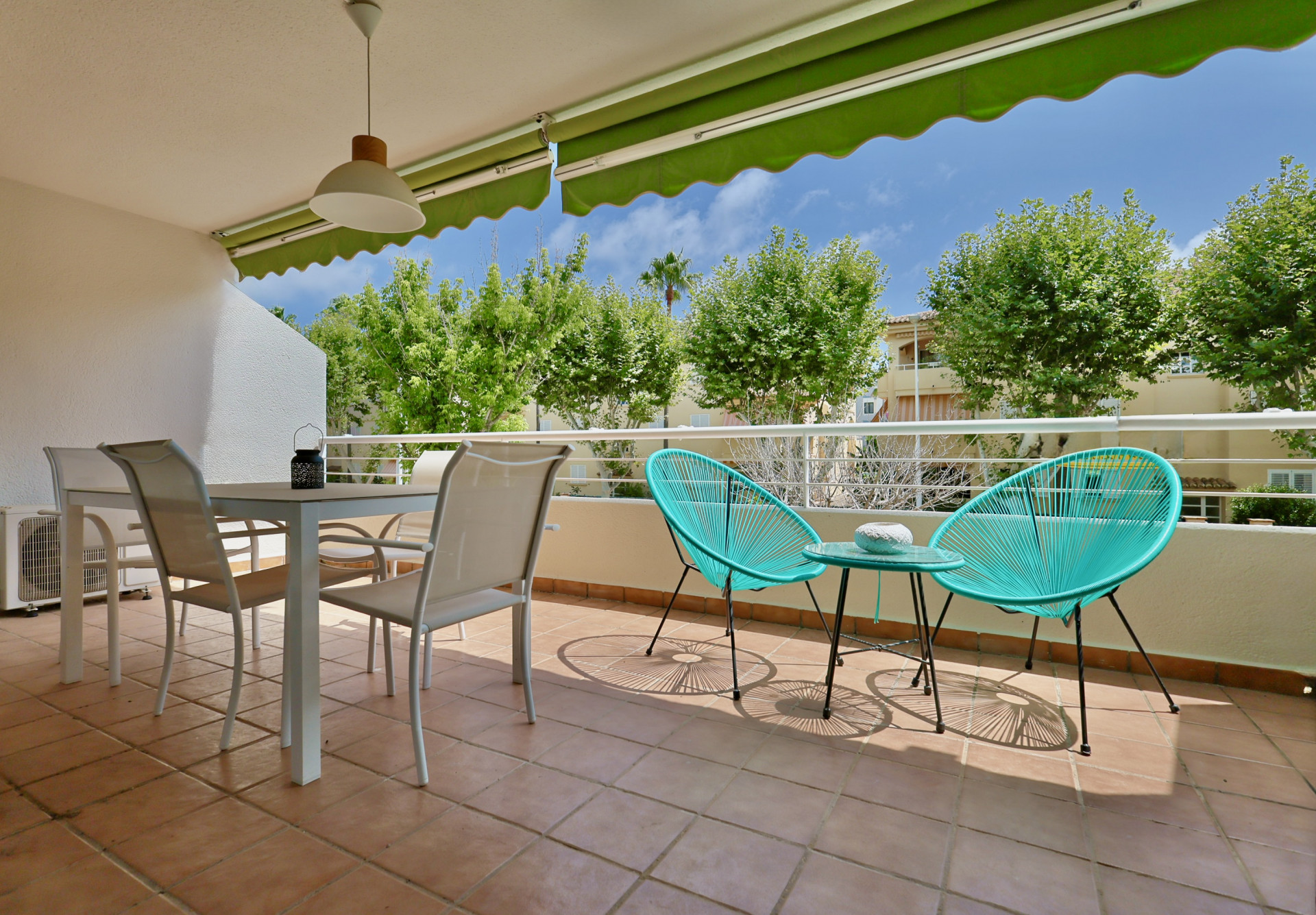Terraza exterior con suelo de baldosas de terracota, conjunto de comedor blanco y sillas Acapulco turquesa bajo un toldo verde extensible.