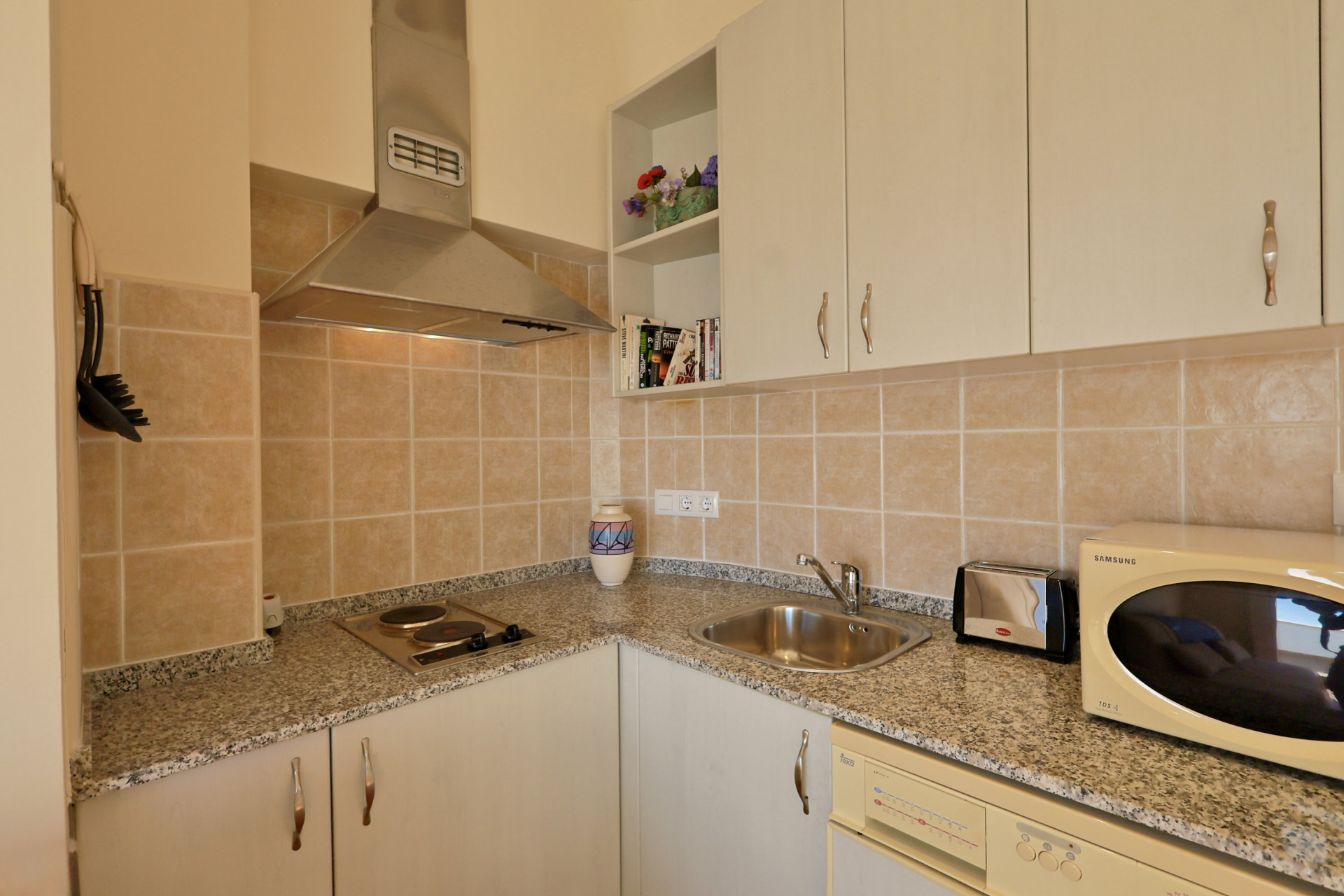 Cocina funcional con encimera de granito moteado, campana de acero inoxidable y azulejos beige. Dispone de armarios blancos y electrodomésticos compactos.