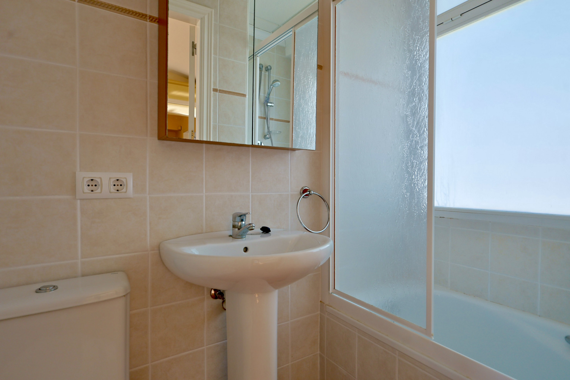 Cuarto de baño con lavabo de pedestal, inodoro y bañera con mampara de cristal translúcido. Revestimiento de azulejos cerámicos en tonos beige.
