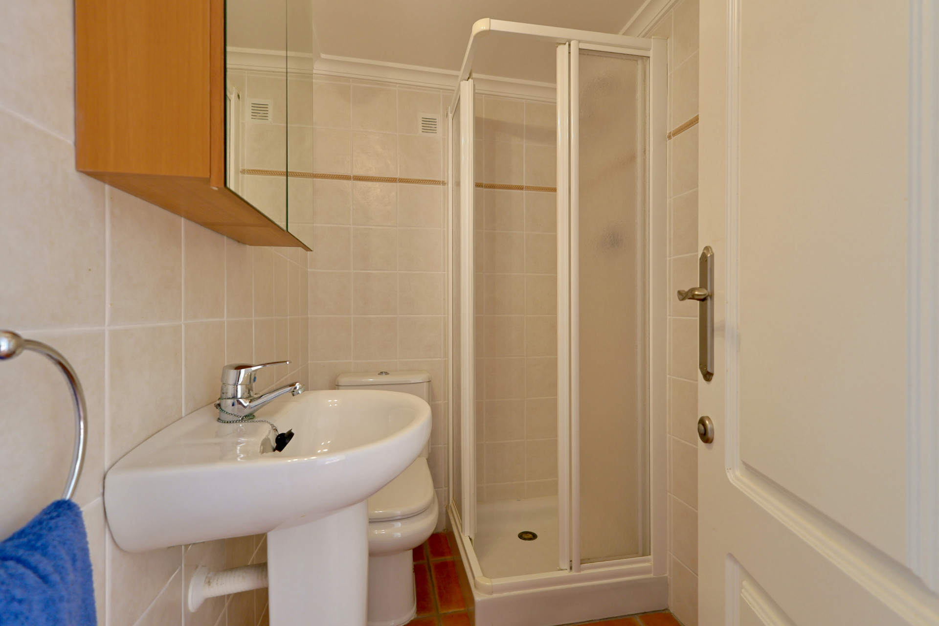 Cuarto de baño funcional con lavabo de pedestal, cabina de ducha de esquina y azulejos en tonos crema con cenefa decorativa.