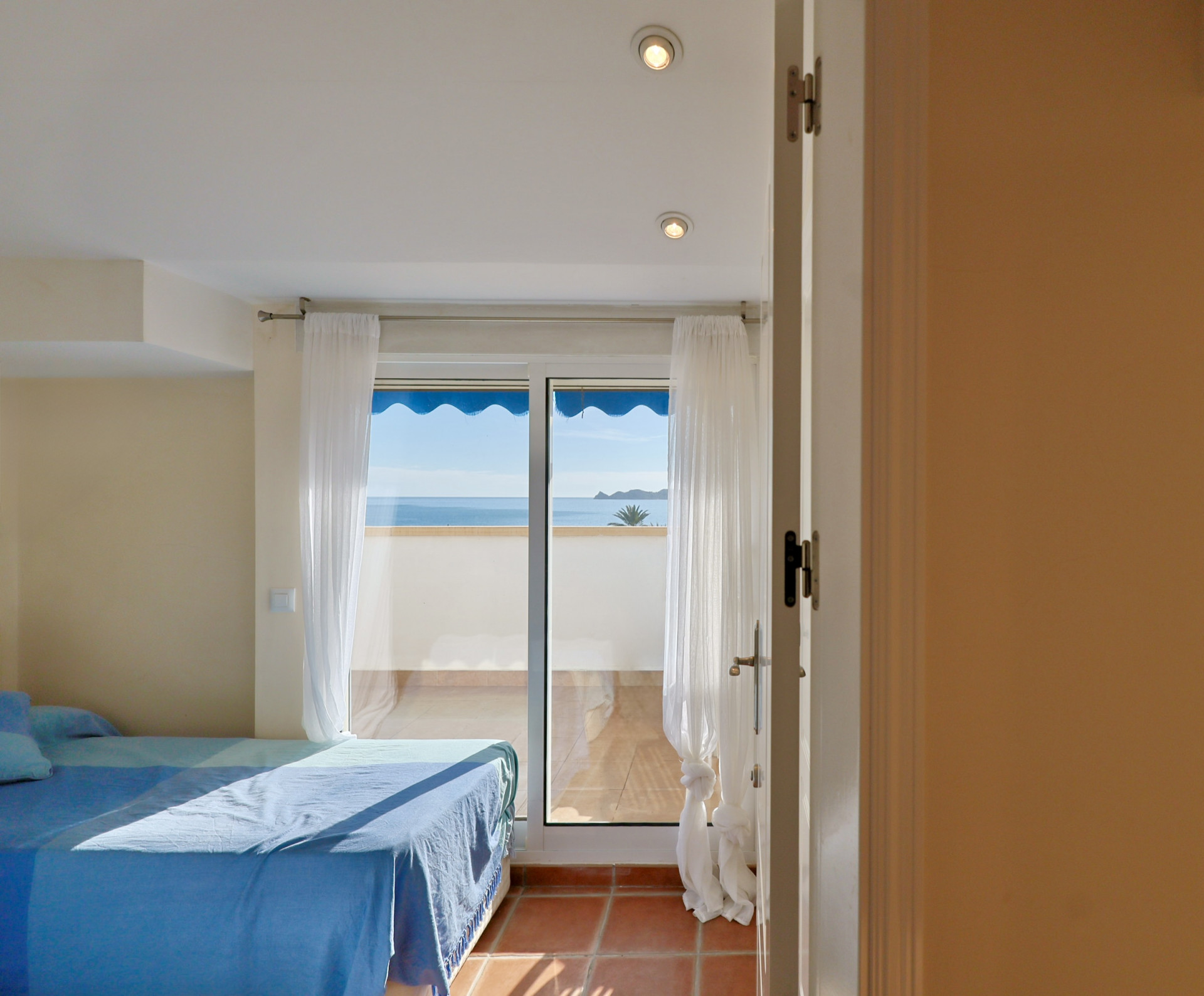 Dormitorio con suelo de terracota y acceso directo a una terraza privada con vistas al mar y toldo azul a través de un ventanal corredero.