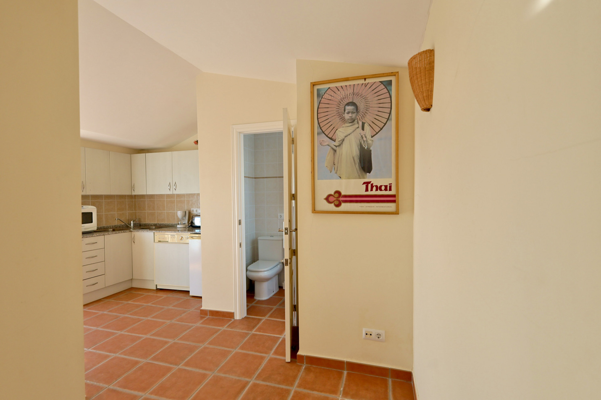 Pasillo con suelos de terracota que conecta con una cocina de armarios blancos y un baño con revestimiento cerámico claro.