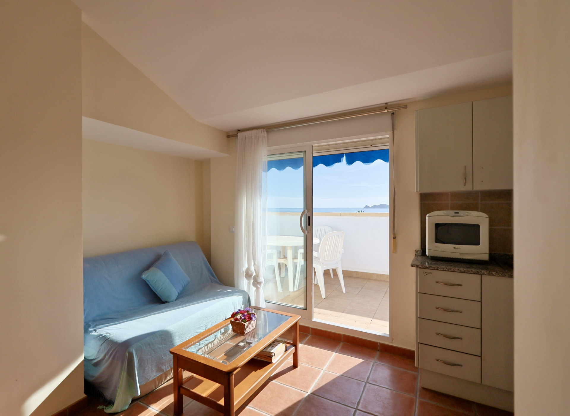 Salón luminoso con suelos de terracota, sofá azul y acceso directo a una terraza privada con vistas al mar y toldo azul.