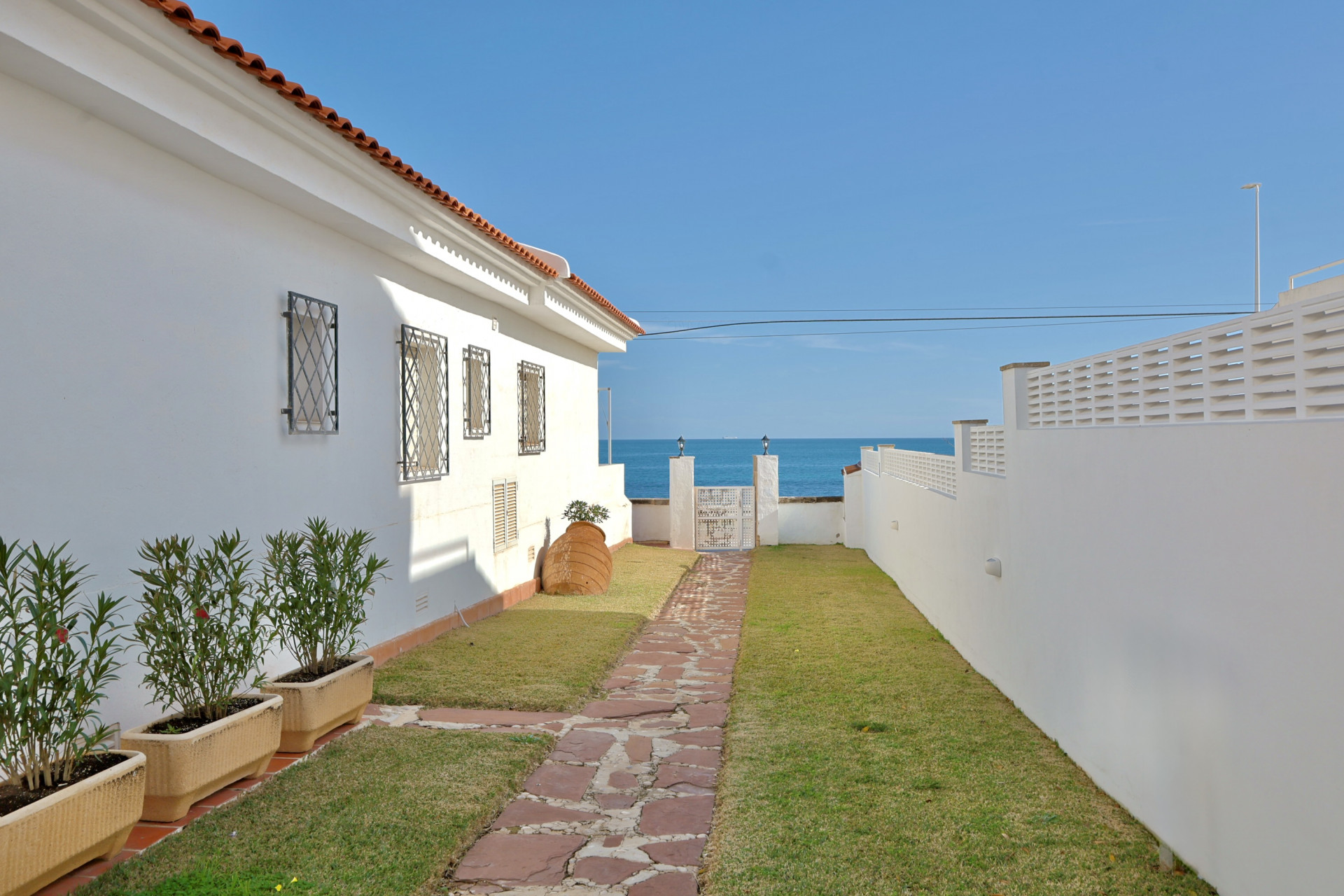 Sendero de piedra natural rodeado de césped que conduce hacia una puerta blanca con vistas despejadas al mar Mediterráneo y al horizonte en Jávea.