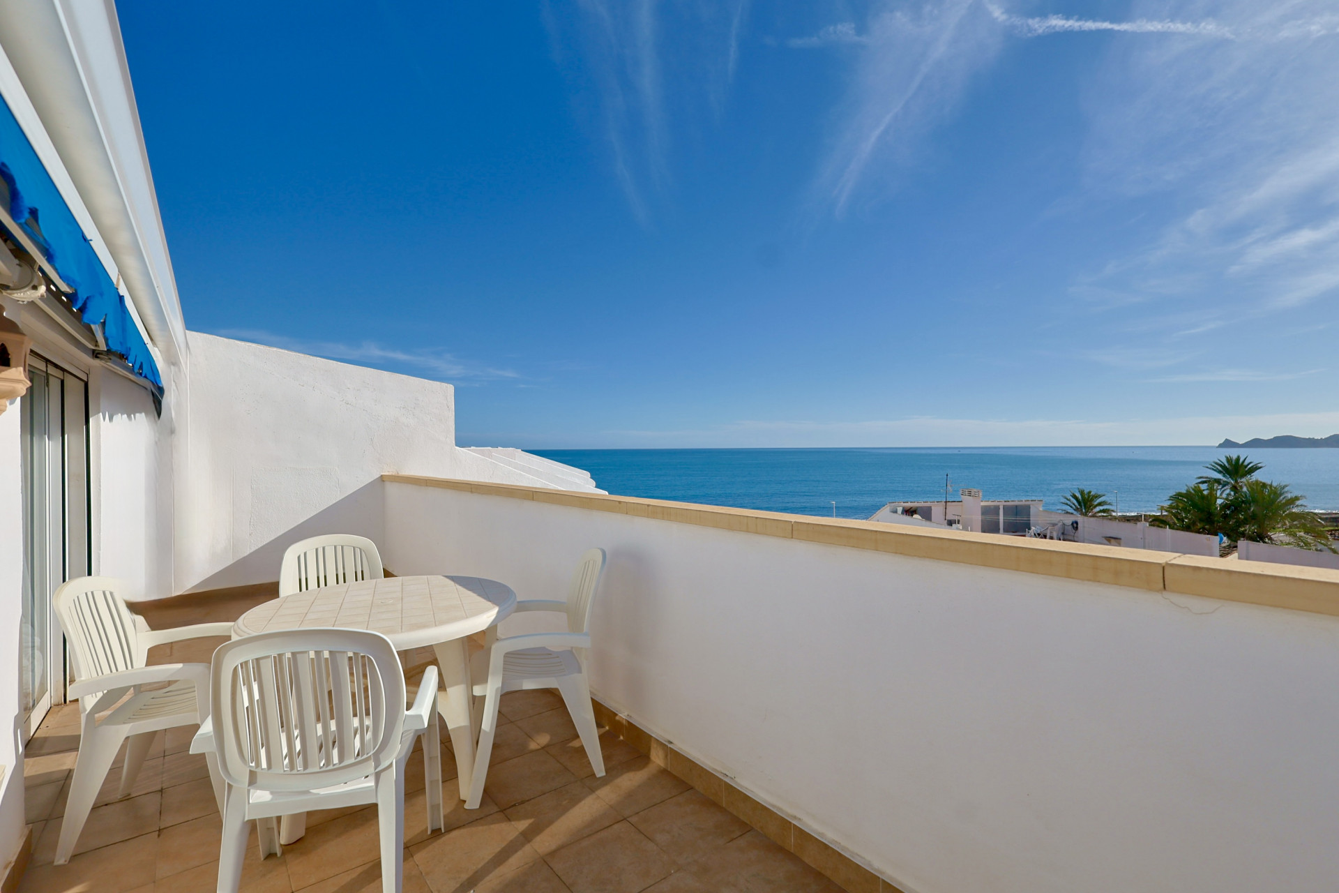Terraza con vistas panorámicas al mar, equipada con mobiliario blanco, suelo cerámico y un toldo azul bajo un cielo despejado.