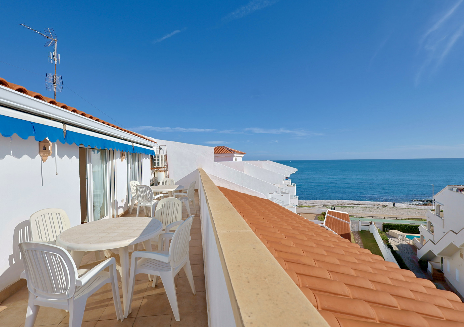 Terraza en primera línea con vistas al mar, equipada con mobiliario de exterior blanco, toldo azul y vistas directas a la playa de grava y el Mediterráneo.