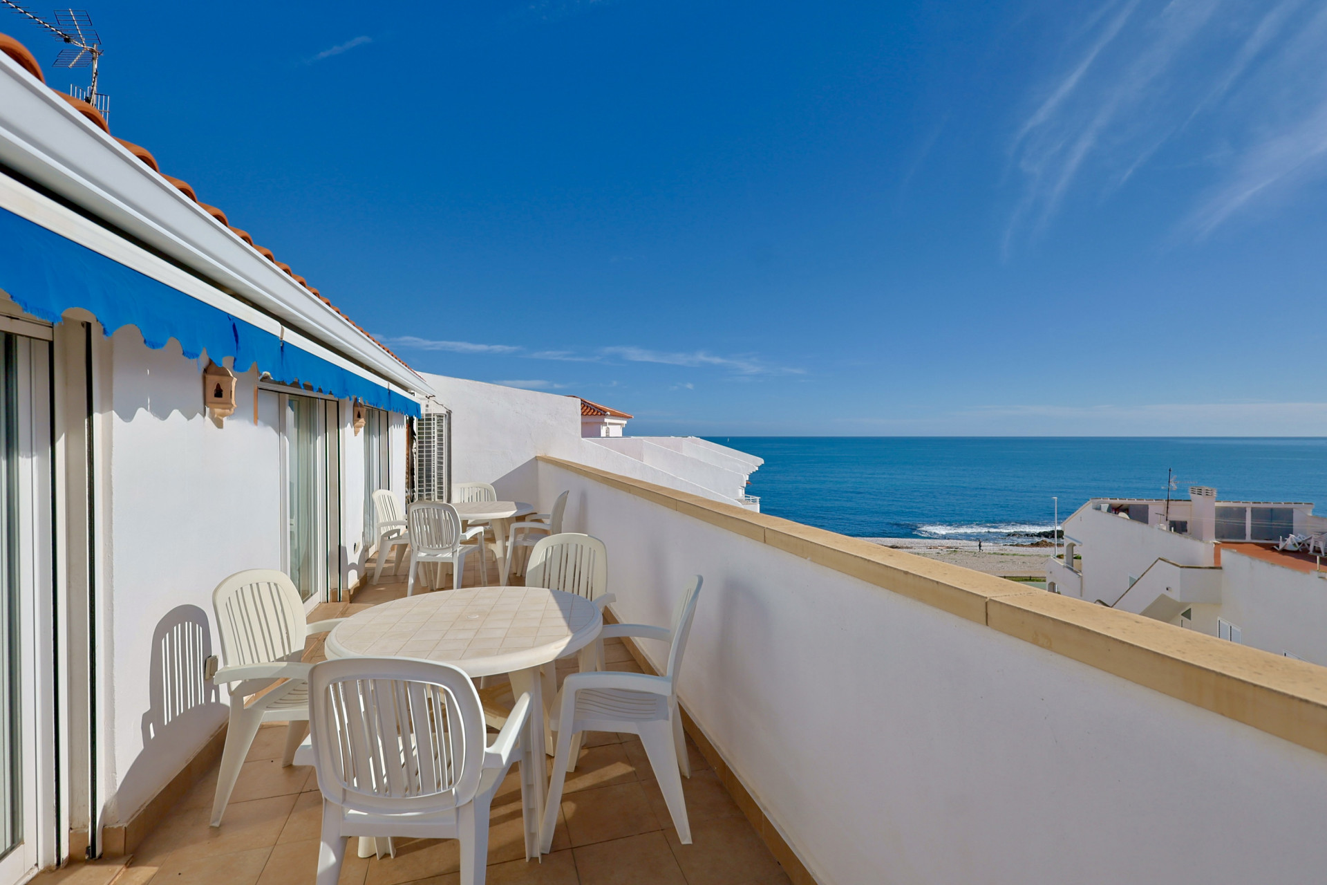 Terraza frontal al mar con pavimentos cerámicos, mobiliario de exterior blanco y toldo retráctil azul con vistas despejadas al Mediterráneo.