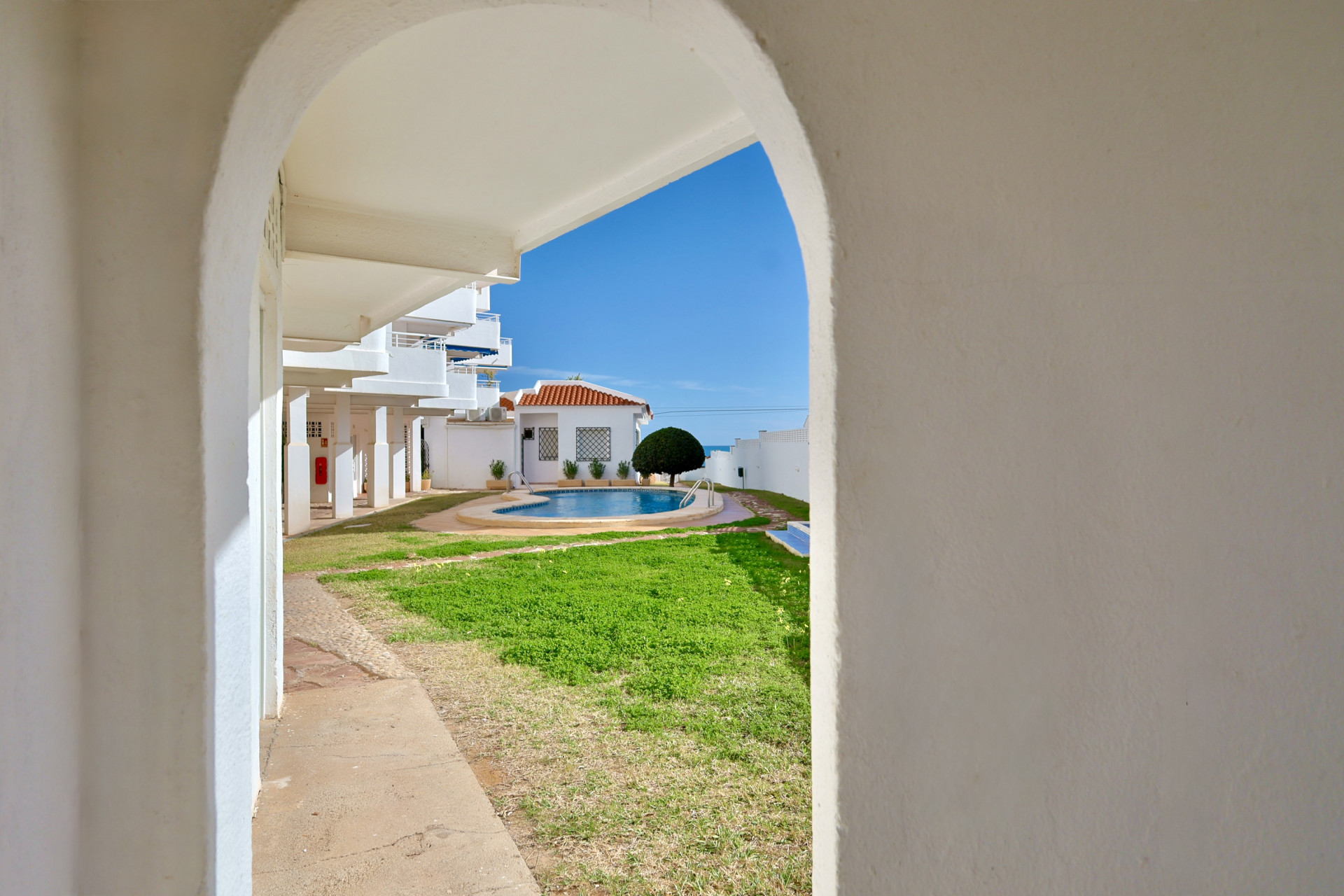 Vista a través de un arco blanco de la piscina comunitaria y zona de césped, rodeada de edificios blancos y bajo un cielo mediterráneo despejado.