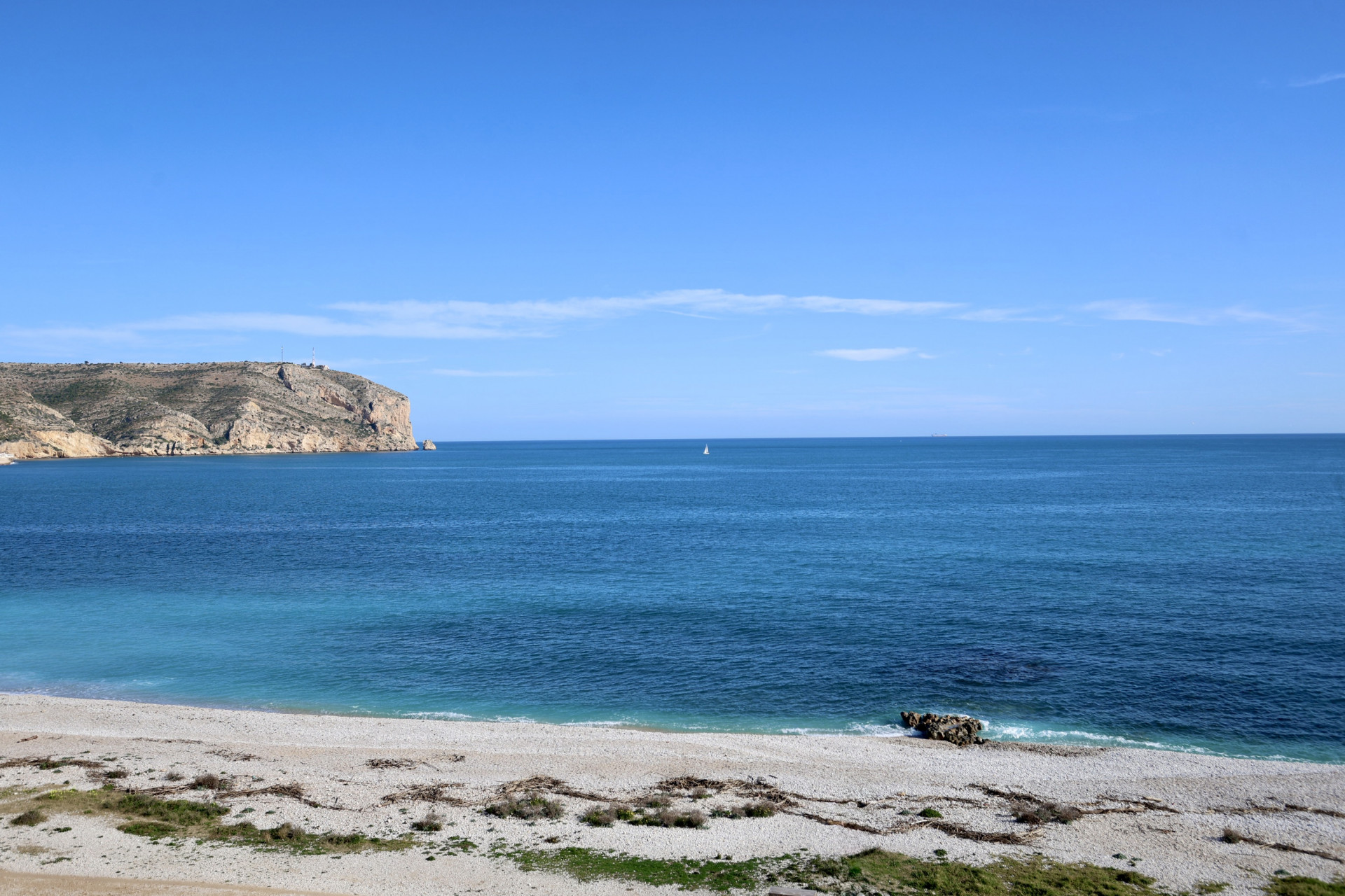 Vista frontal del mar Mediterráneo y el Cabo de San Antonio desde la playa de guijarros en Jávea, bajo un cielo despejado.