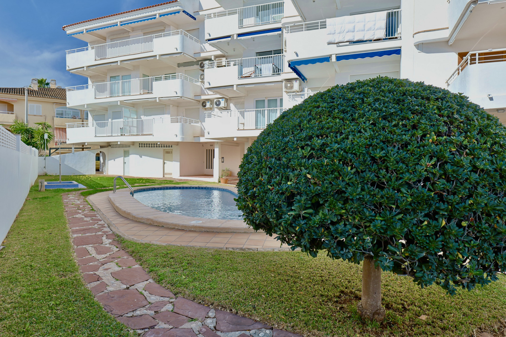 Zona comunitaria con piscina de forma curva, césped cuidado y sendero de piedra natural, situada frente a un edificio blanco con toldos azules.
