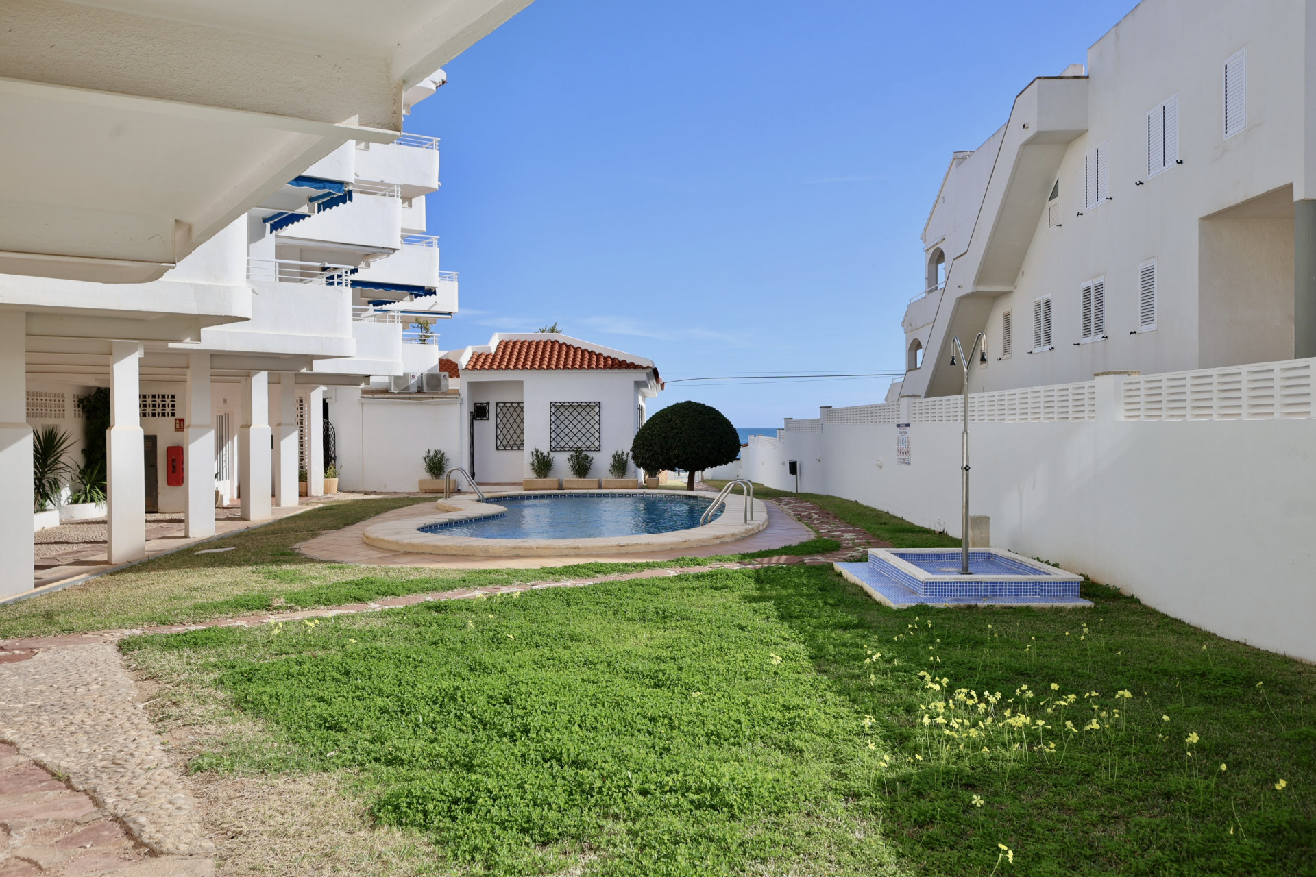 Zona comunitaria con piscina de forma riñonada, jardín de césped y ducha exterior, situada entre edificios blancos con vistas parciales al mar.