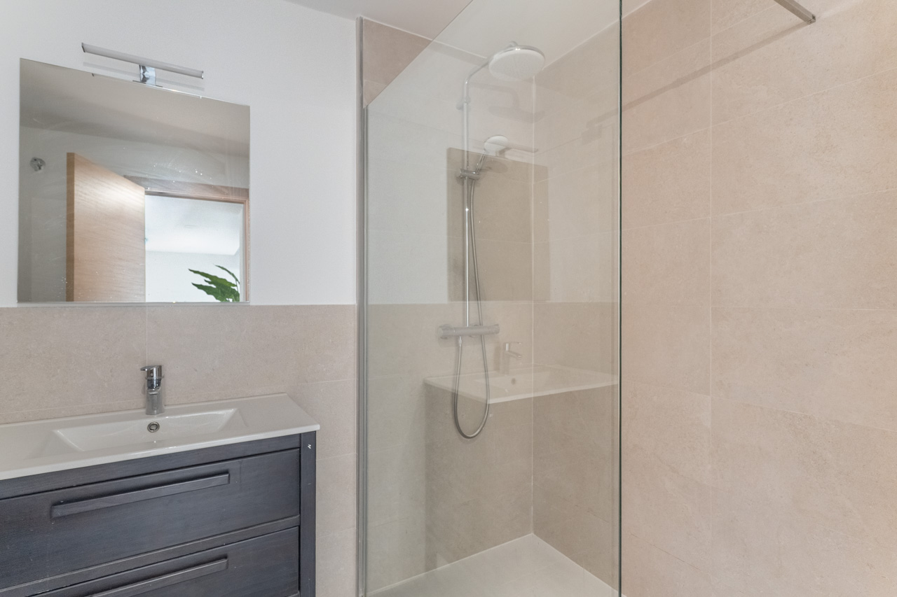 Baño moderno con ducha de obra acristalada, paredes de azulejos beige y mueble de lavabo en madera oscura con espejo cuadrado superior.