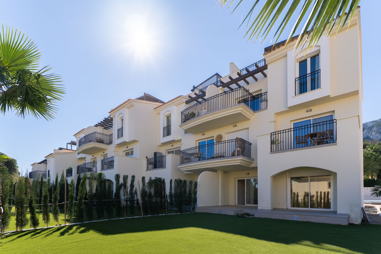 Complejo residencial moderno de fachadas blancas con amplias terrazas, barandillas metálicas negras y zona ajardinada con césped bajo el sol mediterráneo.