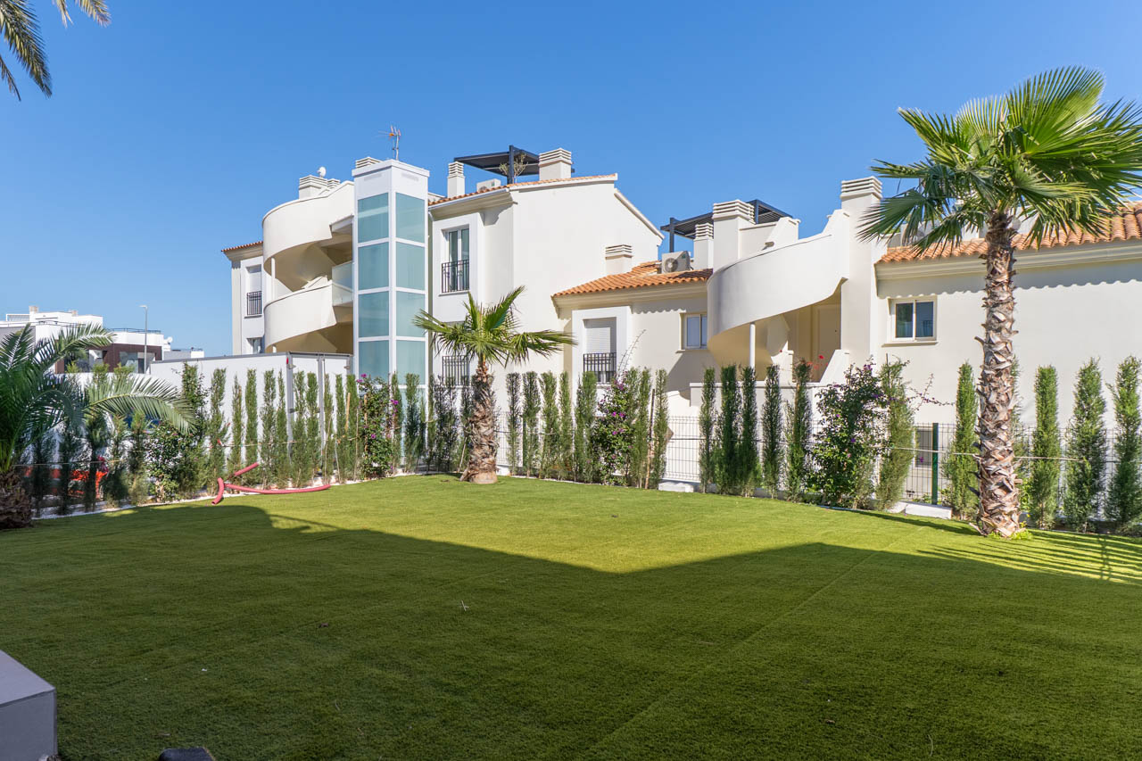 Jardín privado con césped artificial, palmeras y seto perimetral. Destaca la fachada blanca con escaleras de caracol exteriores y un moderno ascensor acristalado.