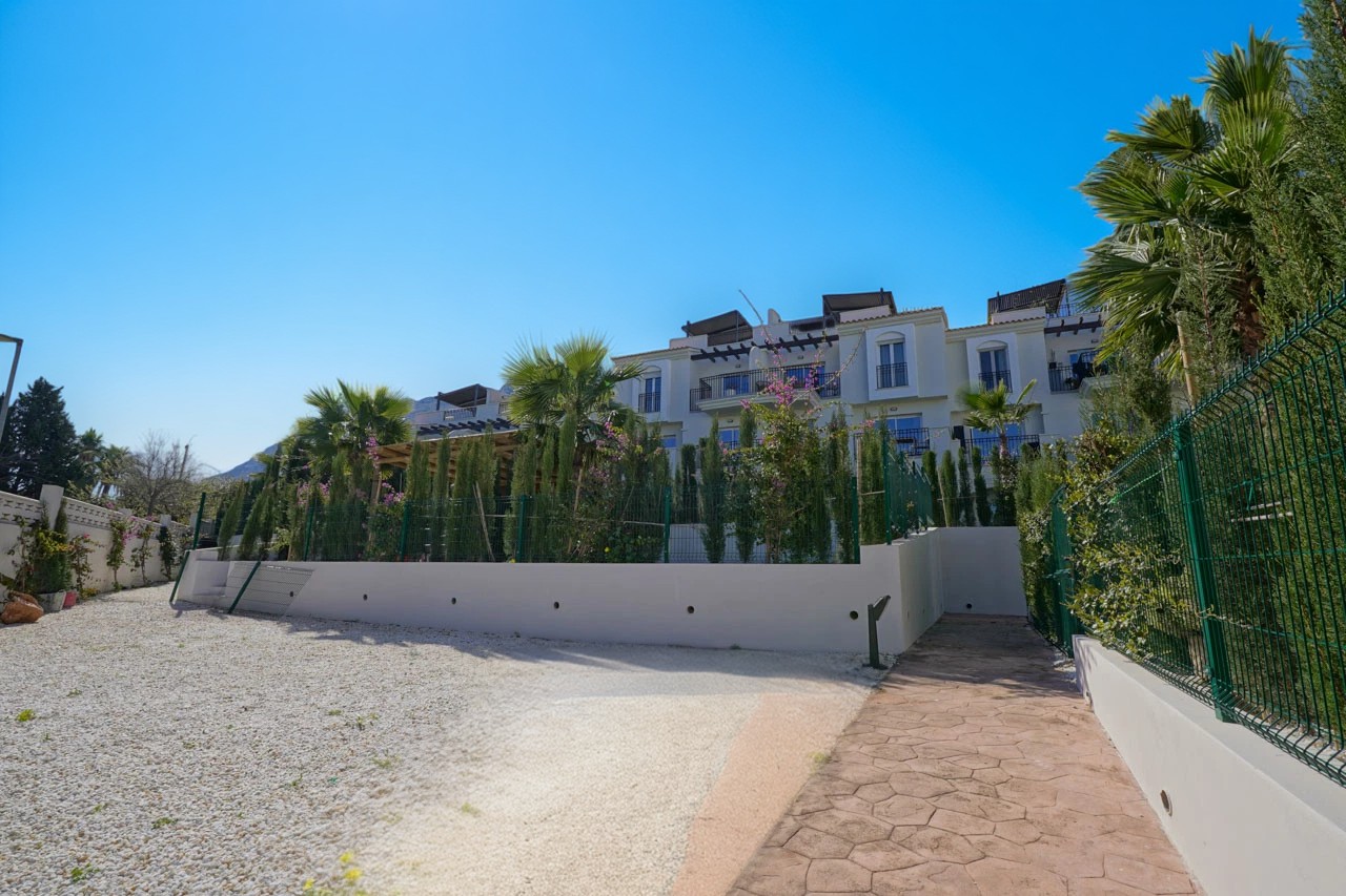 Vista exterior de un complejo residencial blanco con balcones privados, palmeras integradas y un camino pavimentado junto a una zona de grava blanca.