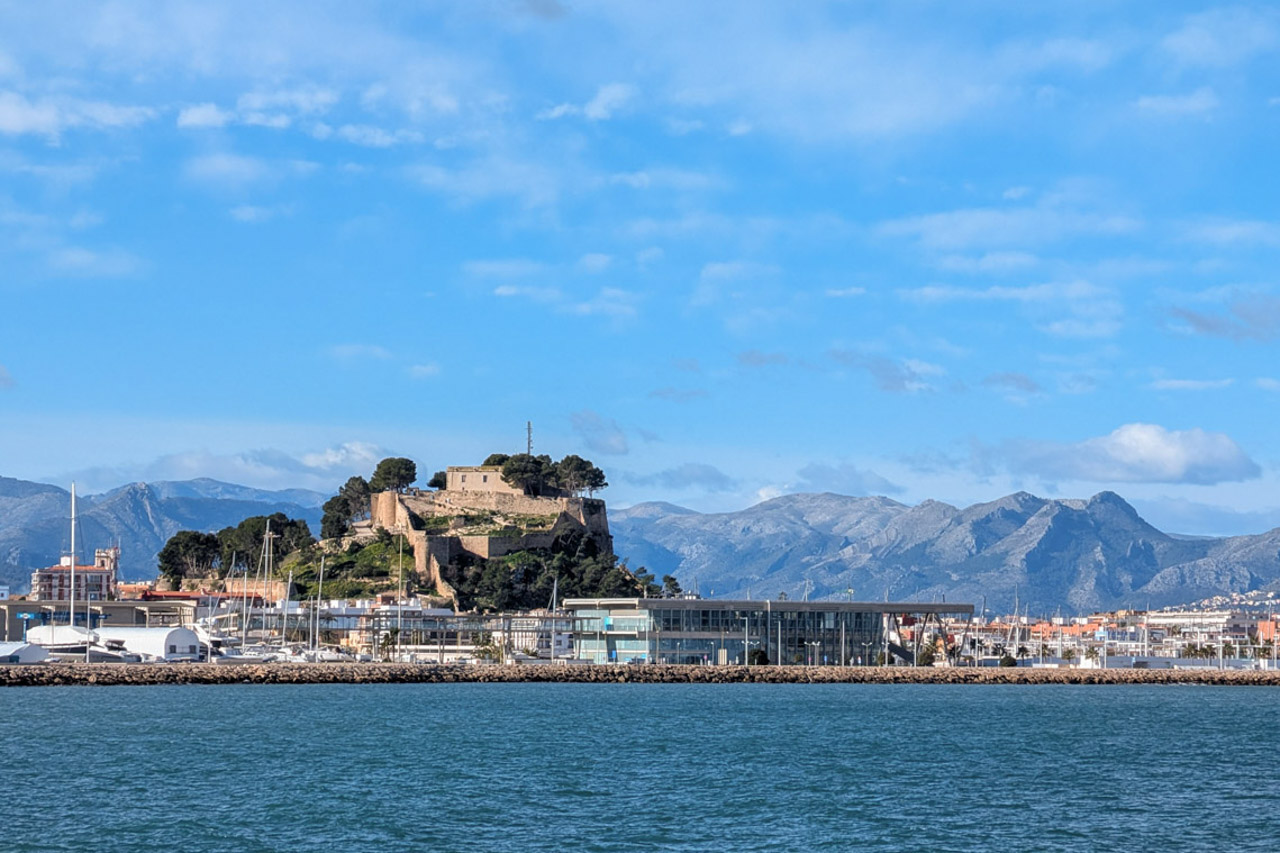 Vistas panorámicas del castillo histórico sobre la colina, el puerto deportivo con instalaciones modernas y las montañas al fondo bajo un cielo despejado.