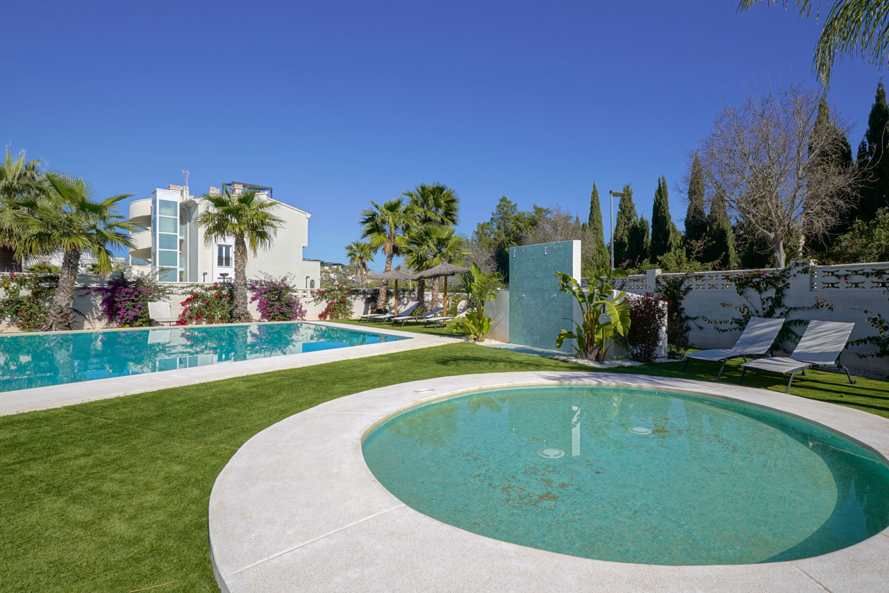 Zona exterior con piscina rectangular, piscina infantil circular, ducha de mosaico turquesa y jardín de césped artificial con palmeras y tumbonas.