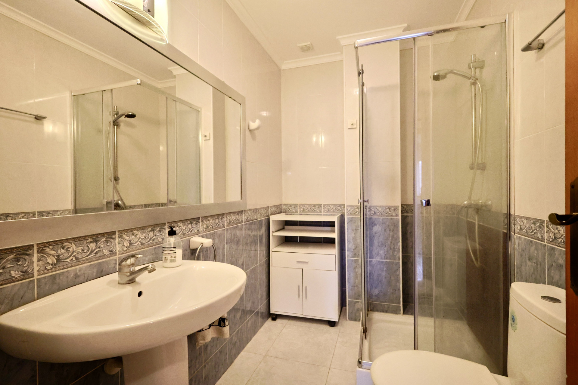 Cuarto de baño con ducha de mampara de cristal, lavabo de pedestal y paredes alicatadas en tonos azules con cenefa decorativa.