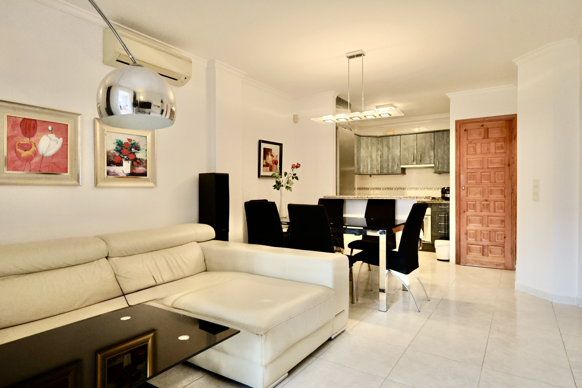 Espacio diáfano con sofá de cuero blanco, mesa de comedor de cristal negro y cocina integrada con armarios grises y encimera de granito.