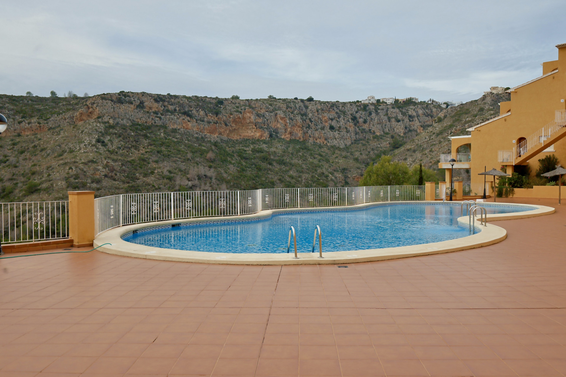 Piscina comunitaria de diseño curvo con amplia terraza de baldosas de terracota y vistas despejadas al valle montañoso de Benitachell.