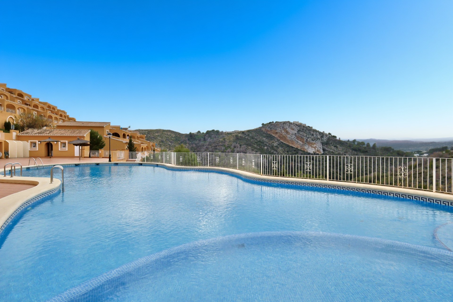 Piscina comunitaria de forma curva con gresite azul y valla de seguridad blanca, con vistas despejadas al valle y las montañas de Benitachell.