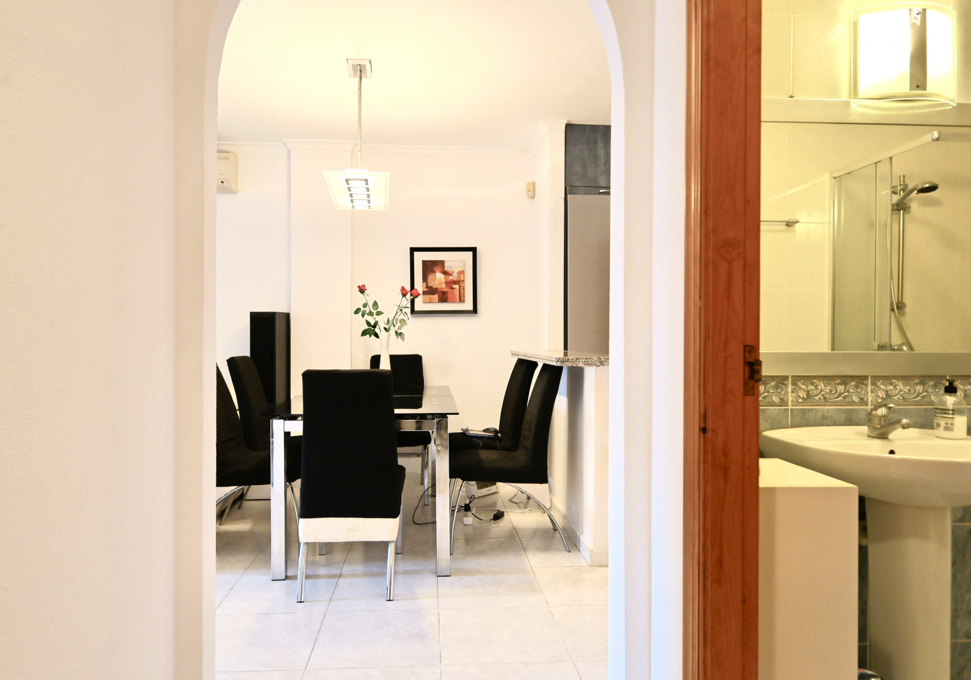 Vista a través de un arco hacia el comedor con mesa de cristal y sillas negras, junto a un baño con lavabo de pedestal y azulejos decorativos.