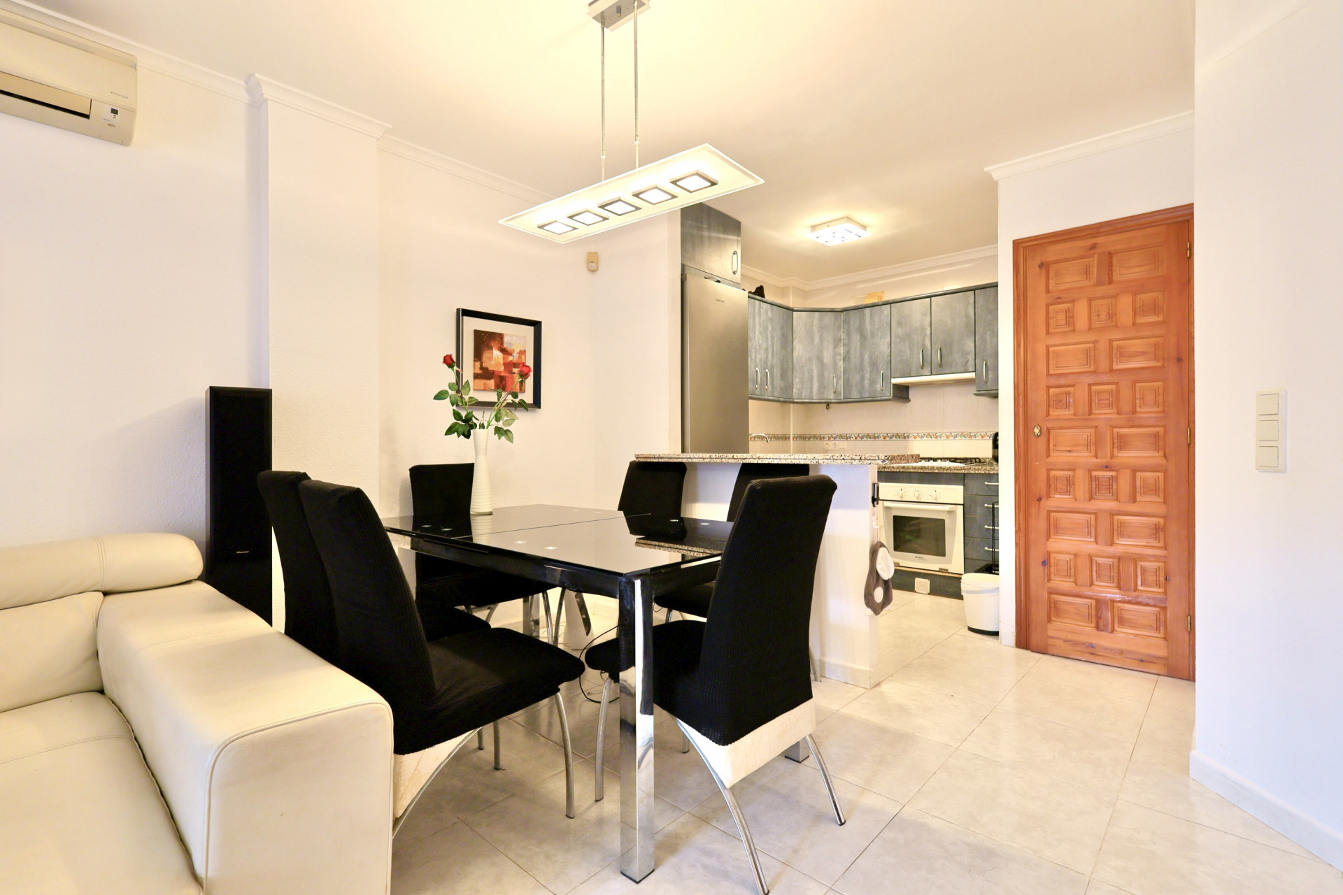 Zona de estar de planta abierta con mesa de comedor de cristal negro, cocina integrada con armarios azules y encimeras de granito, y suelos de gres claro.