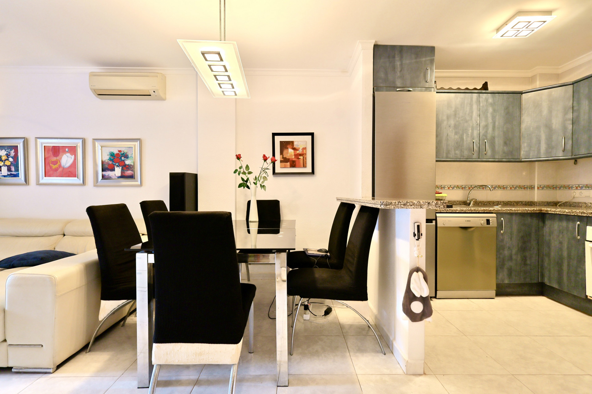 Zona de estar de planta abierta con un comedor de mesa de cristal y sillas negras, junto a una cocina con armarios azules y encimera de granito.