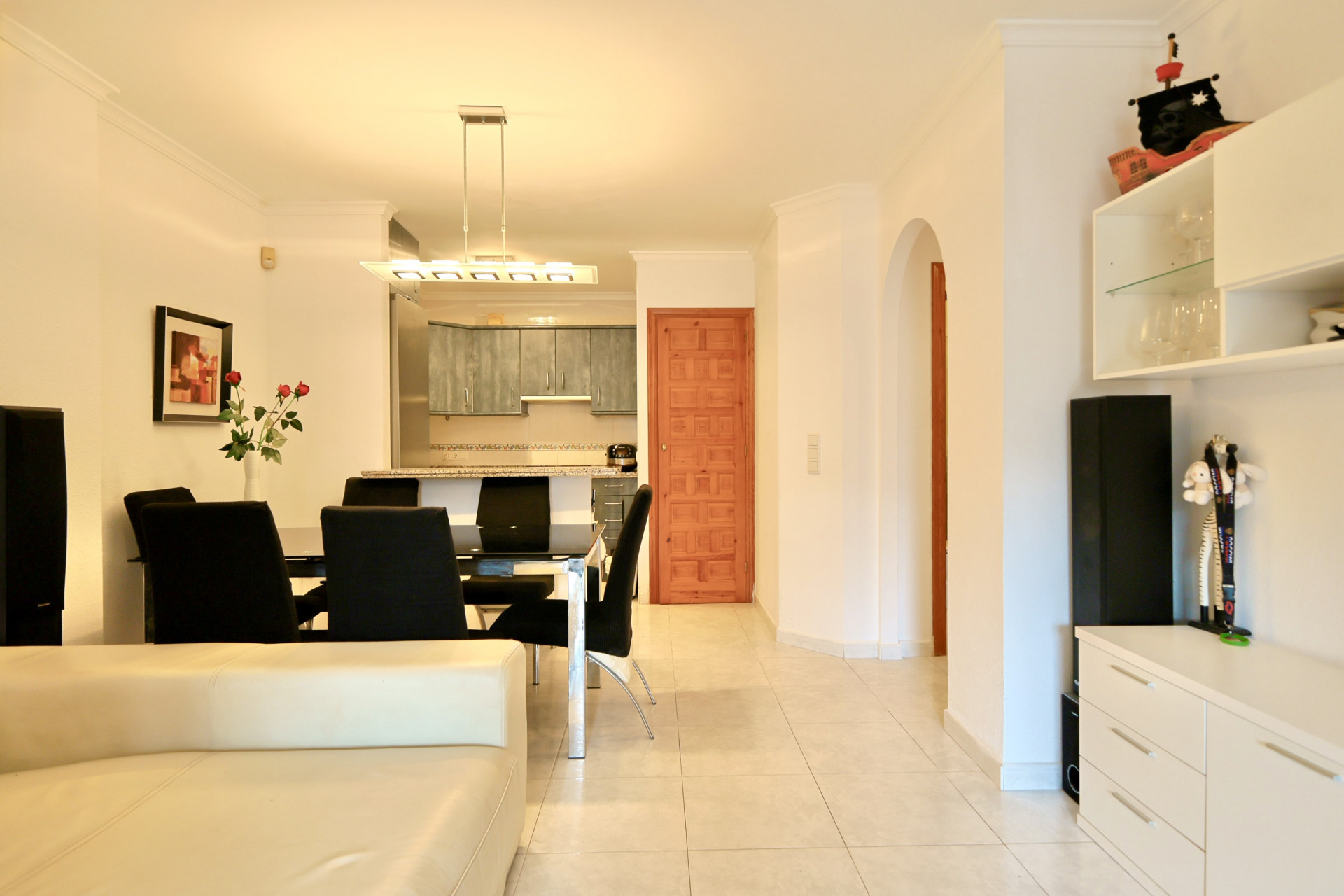 Zona de estar diáfana con comedor de sillas negras, lámpara de diseño y cocina gris al fondo. Suelo de baldosas claras, puerta de madera y arco de paso.